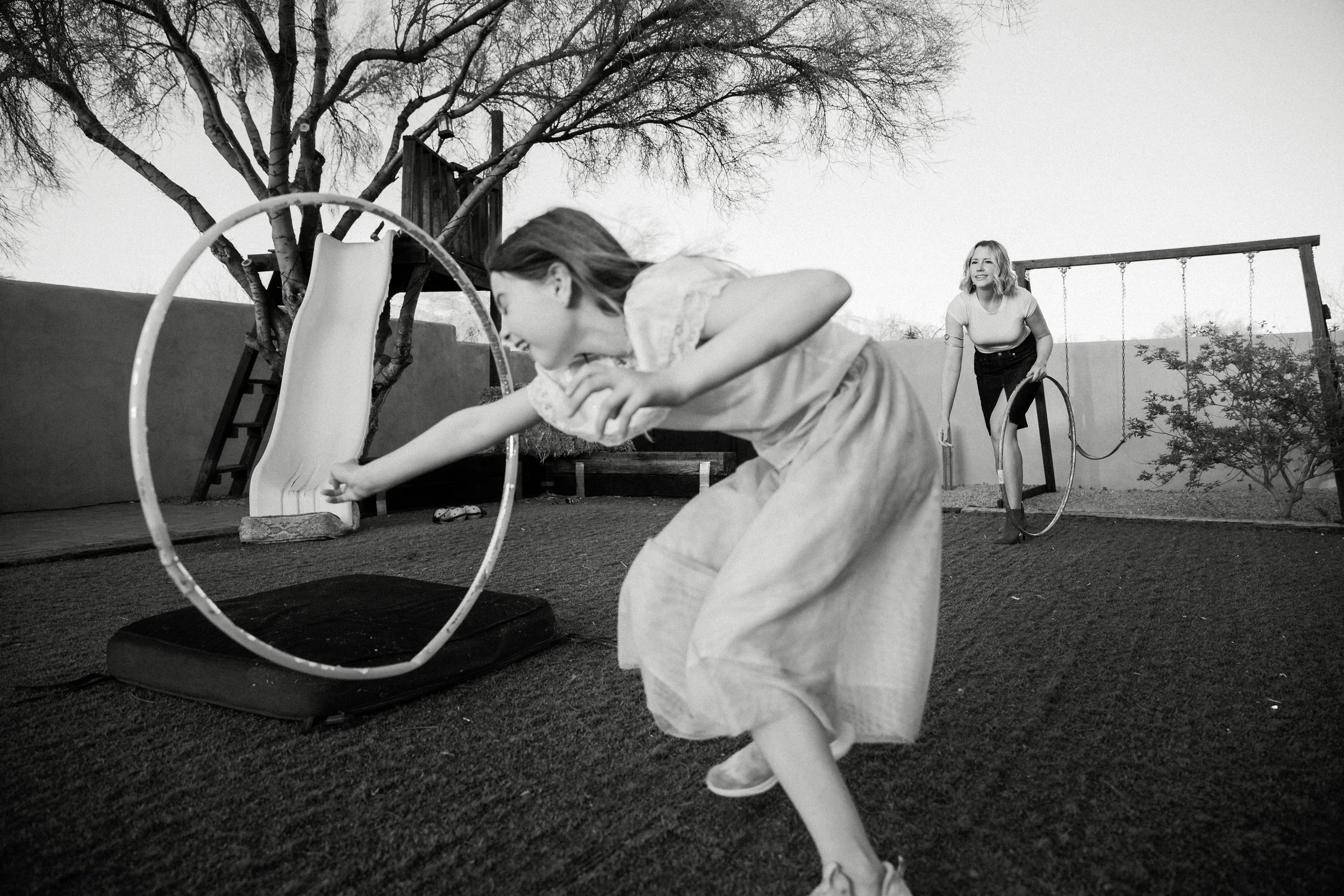 Two children playing with hula hoops in a backyard playground with a slide and swings, some trees and a wall in the background.
