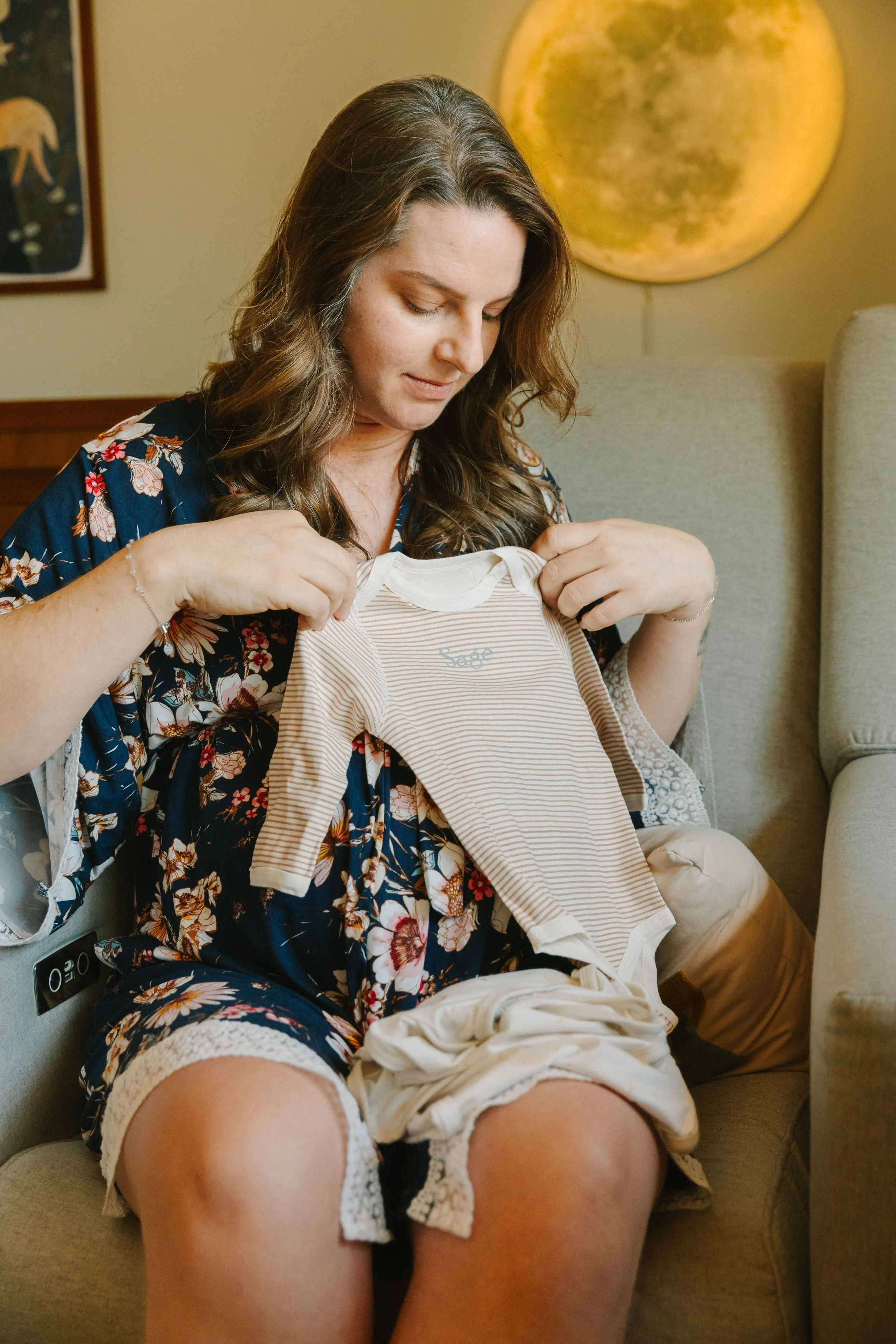 A woman with brown hair and a floral dress is sitting on a beige chair holding a small baby onesie, with a moon painting on the wall behind her.