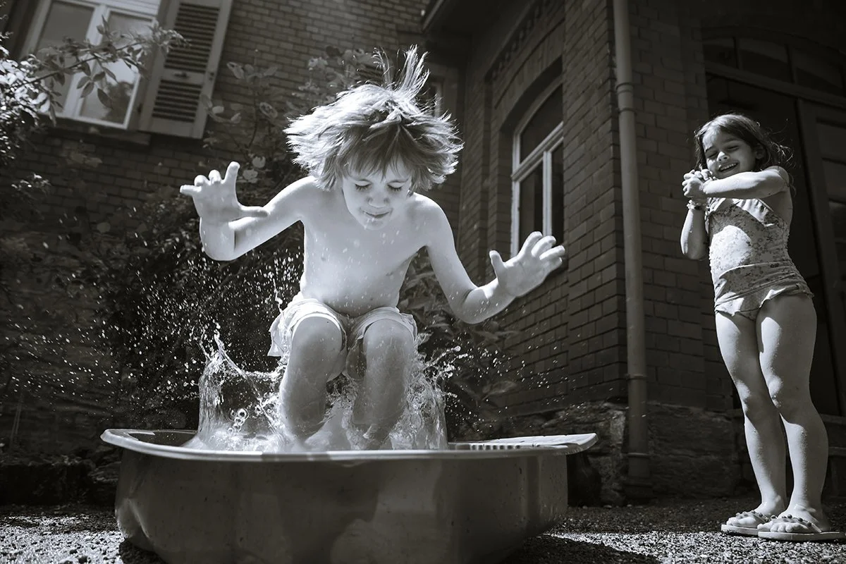 Child jumping into a plastic kiddie pool with water splashing, while another girl watches and laughs nearby outside a brick house.