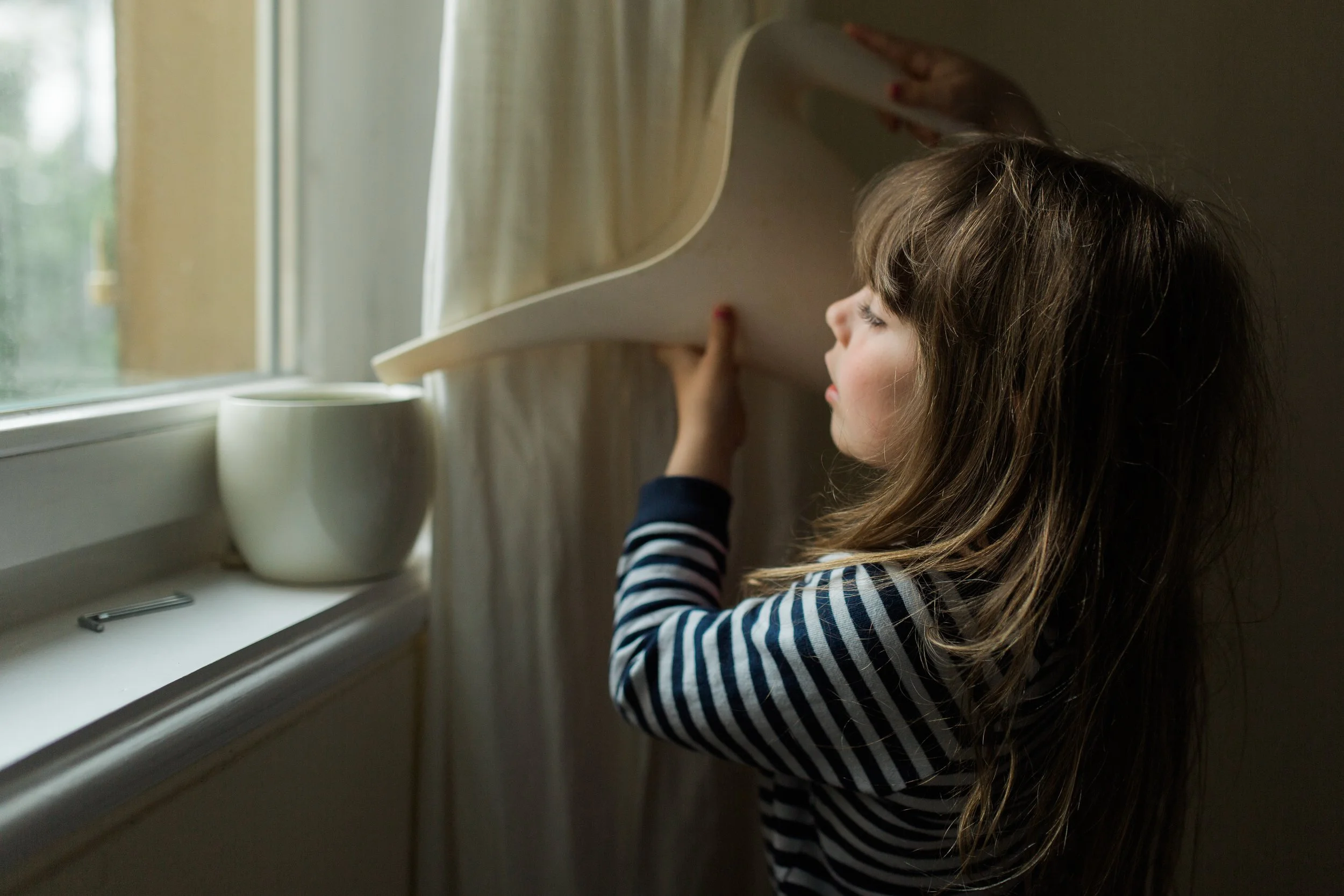 A young girl with brown hair and a striped shirt watering a plant in a window.