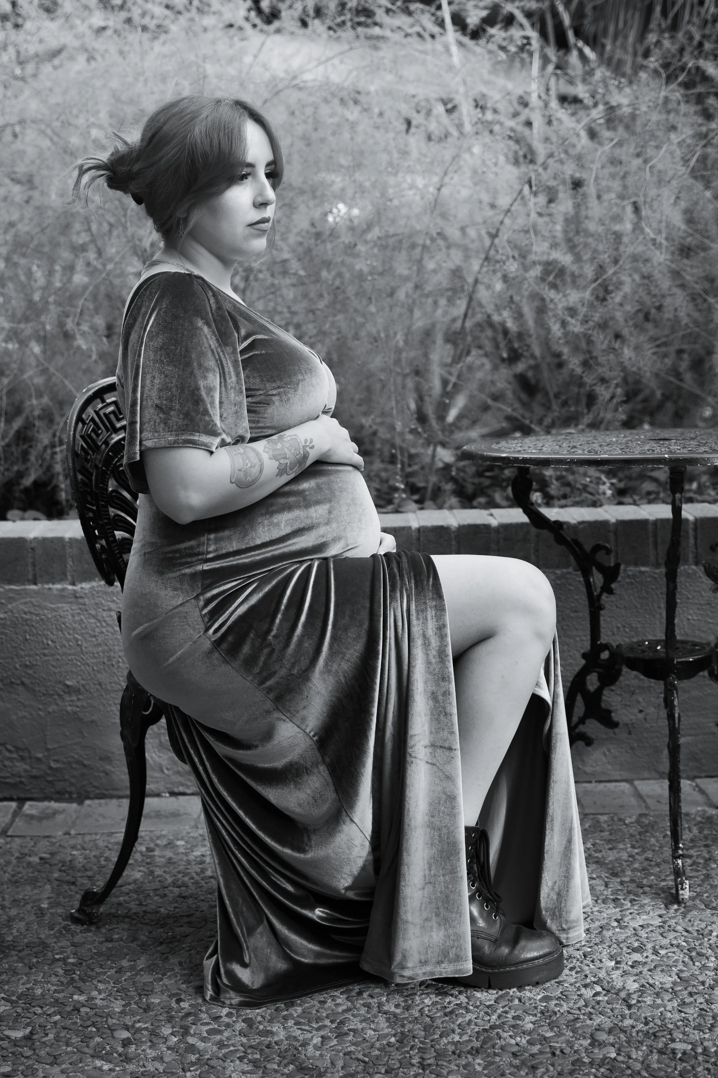 A black and white photo of a woman sitting on a decorative metal chair outdoors. She has shoulder-length hair tied back, tattoos on her forearm, and is wearing a velvet top, a long skirt, and combat boots. She looks to the side with a contemplative expression.
