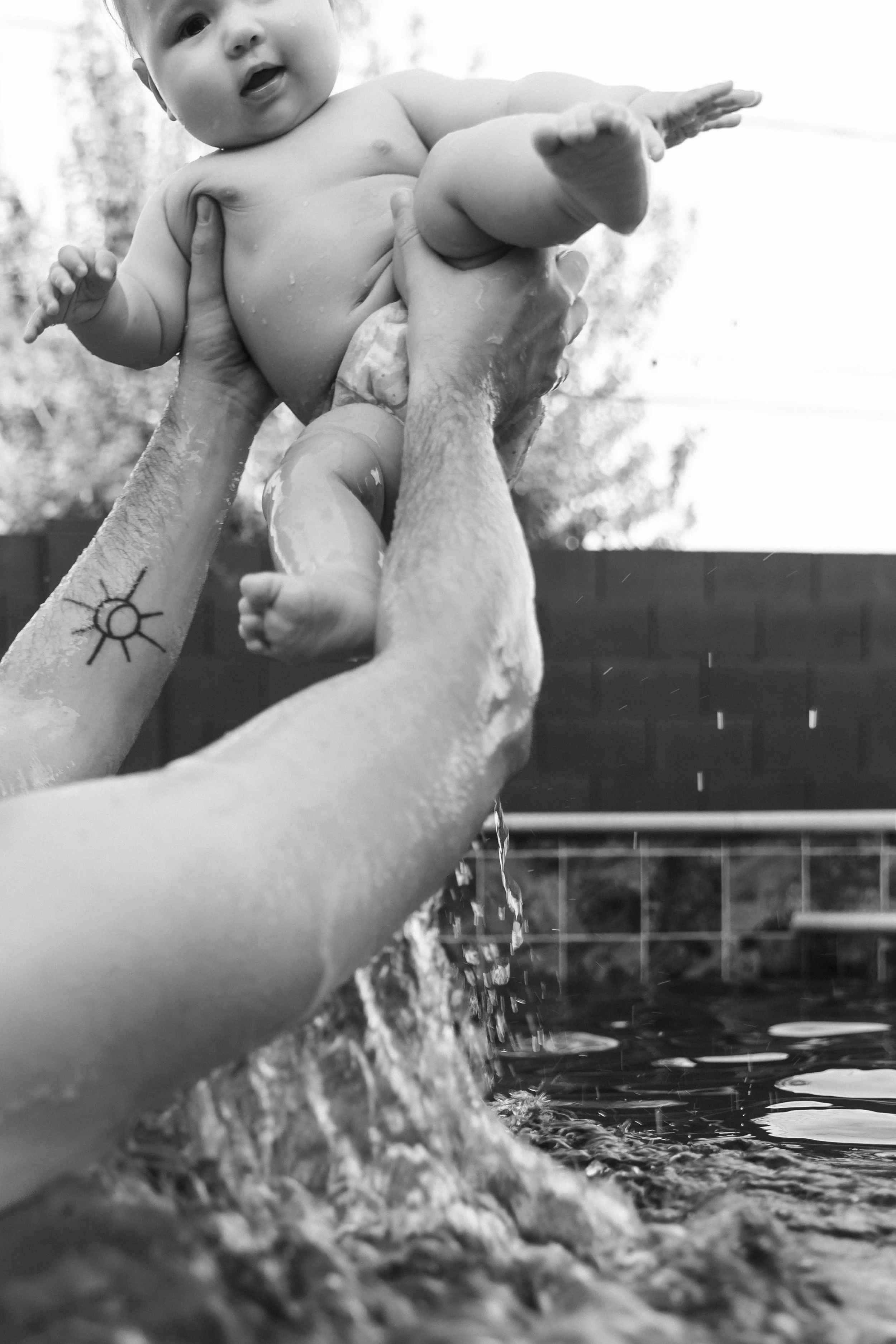 A black and white photo of a baby being lifted out of water by two adult hands, with water dripping from the baby and a tattoo of a sun on one arm.