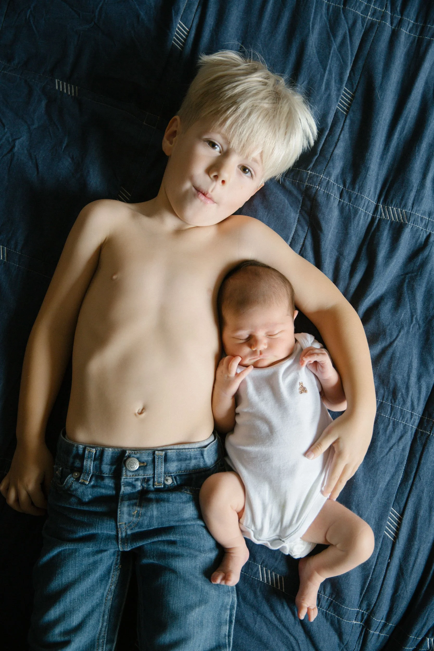 A young boy with blond hair lying shirtless on a bed with a newborn baby. The boy has a relaxed expression, and the baby is asleep, wearing a white onesie.