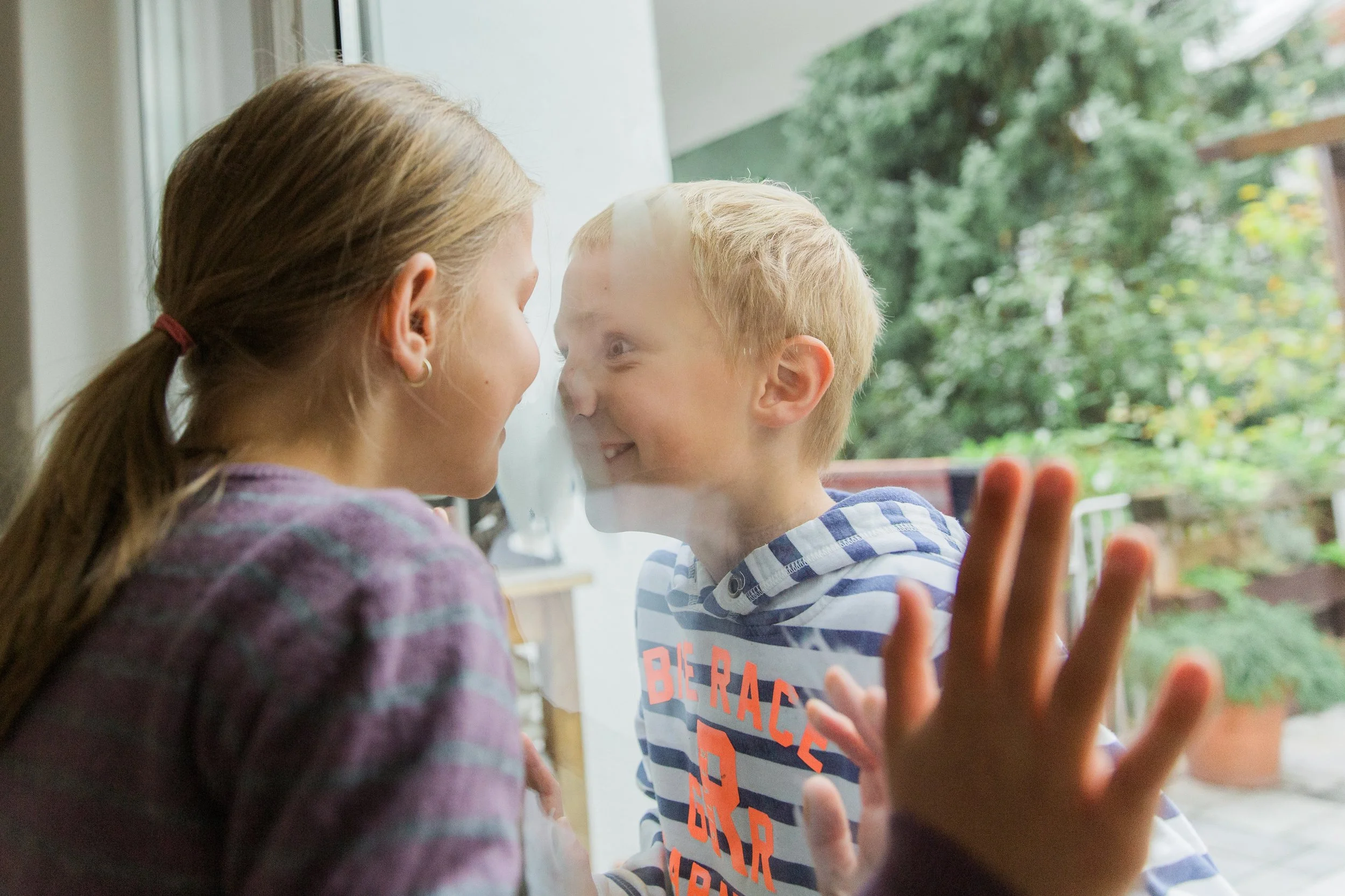 A young girl and a young boy are standing close to each other inside, looking into each other's eyes through a glass door, with the girl pressing her hand against the glass and the boy smiling.