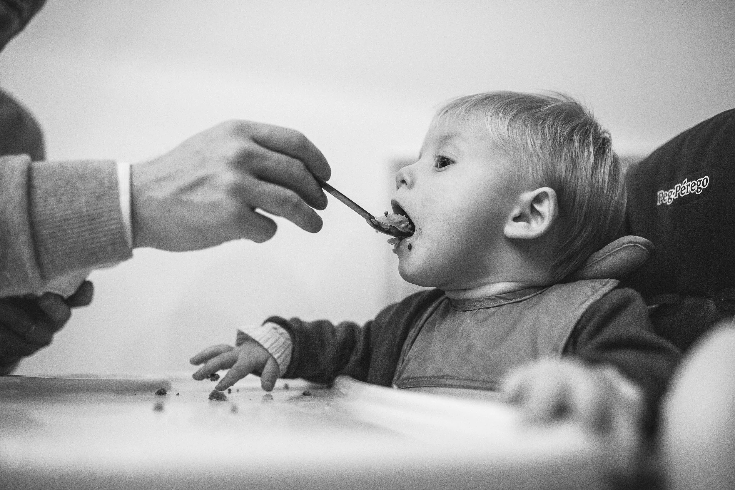 A young boy sitting at a table with his mouth open, being fed with a spoon by an adult hand, with some crumbs on the table and the boy wearing a bib.