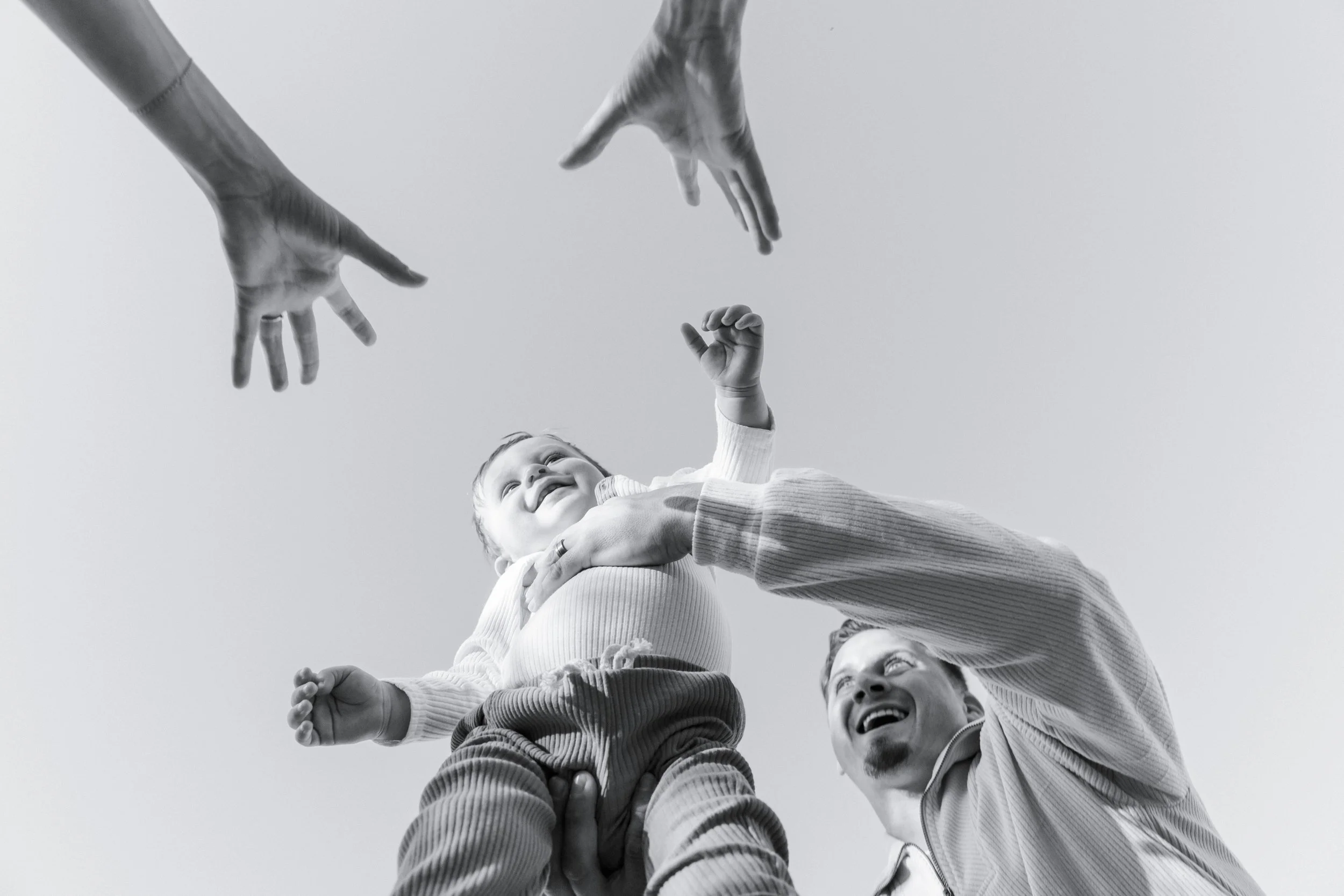 A happy baby being held up in the air by an adult, with two other adults reaching towards the baby in a playful manner.