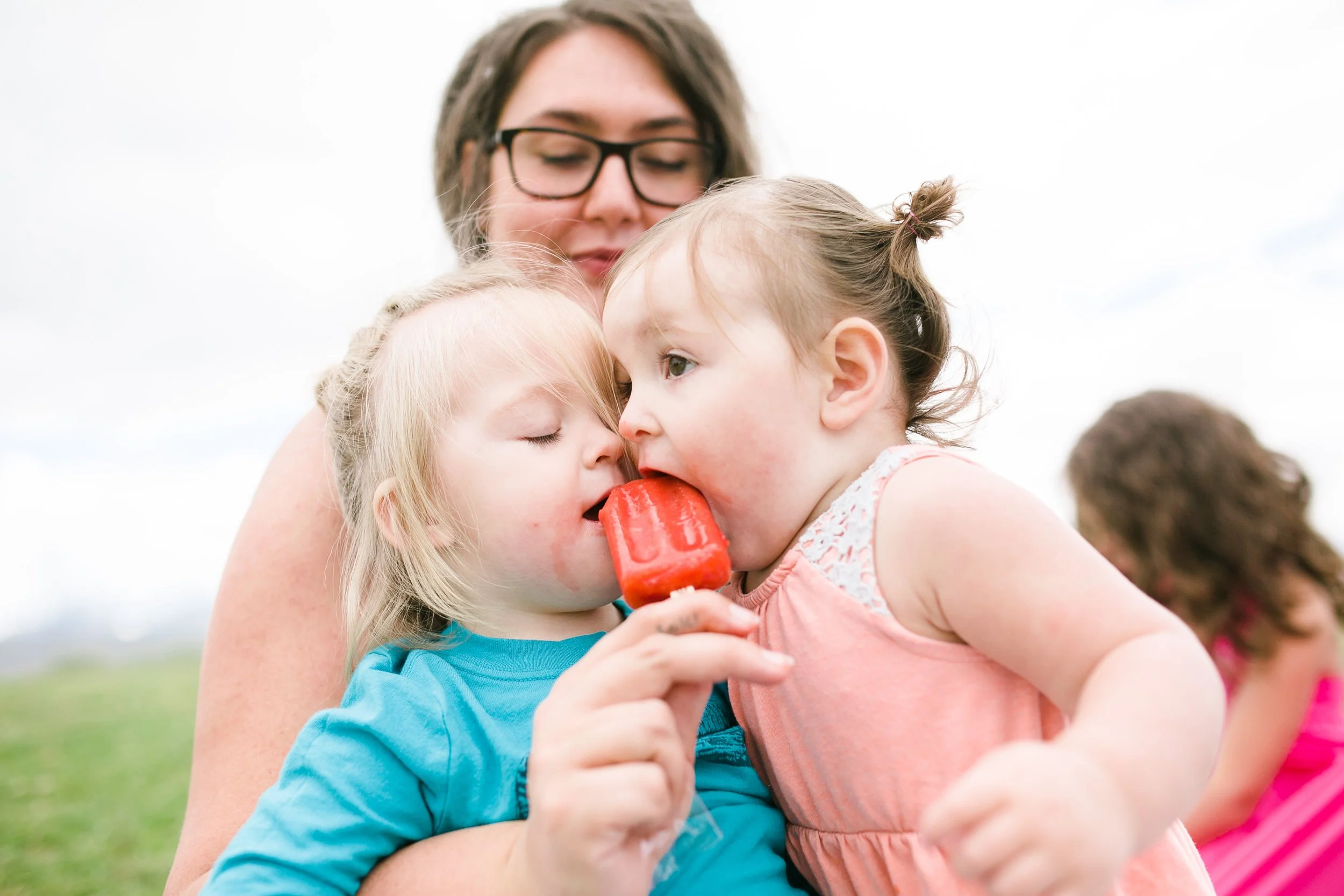 A woman holding two young children, one with blonde hair and the other with brown hair, sharing a red Popsicle outdoors under a cloudy sky.