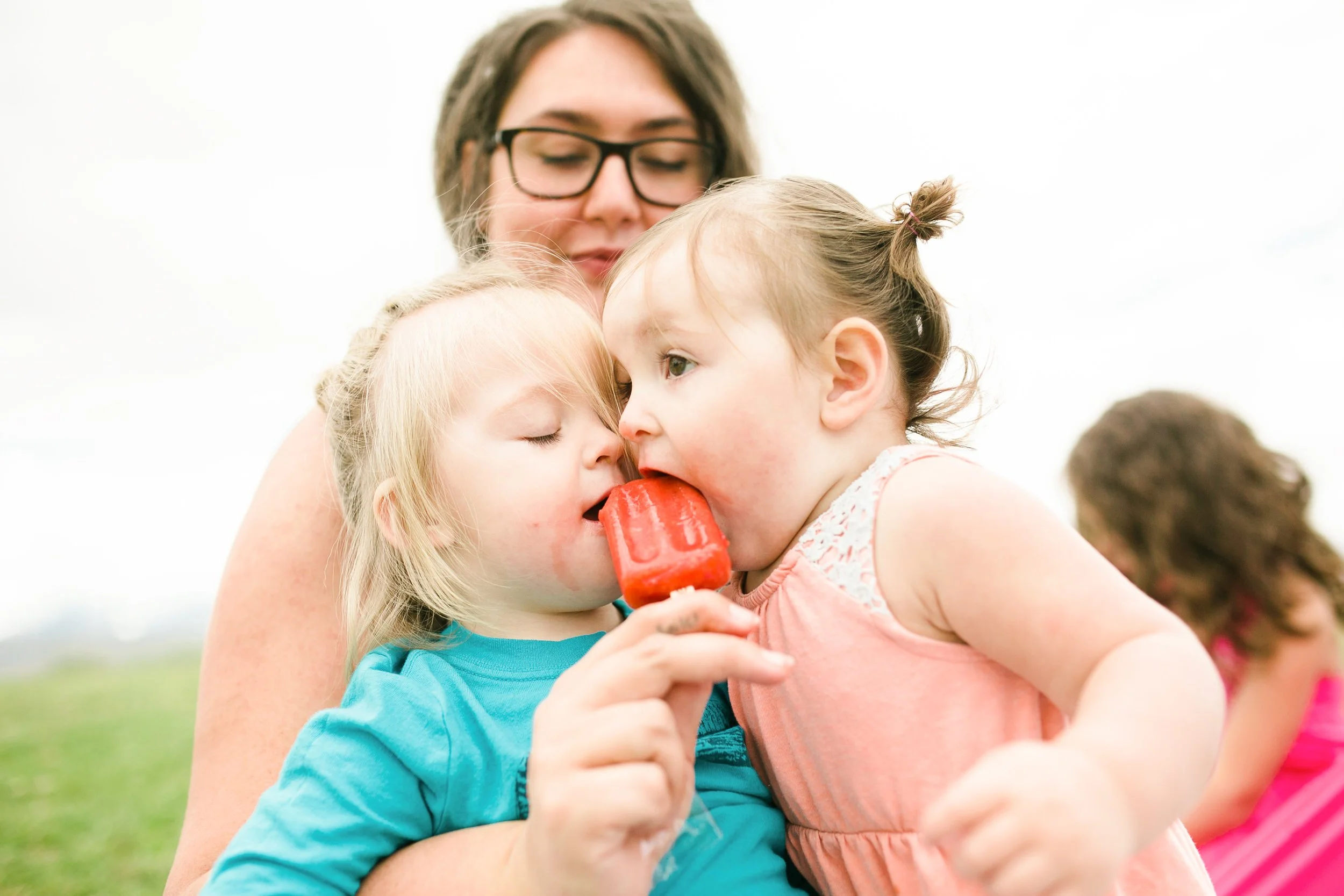 A woman and twin siblings sharing a close moment outdoors, with the twins eating red popsicles.