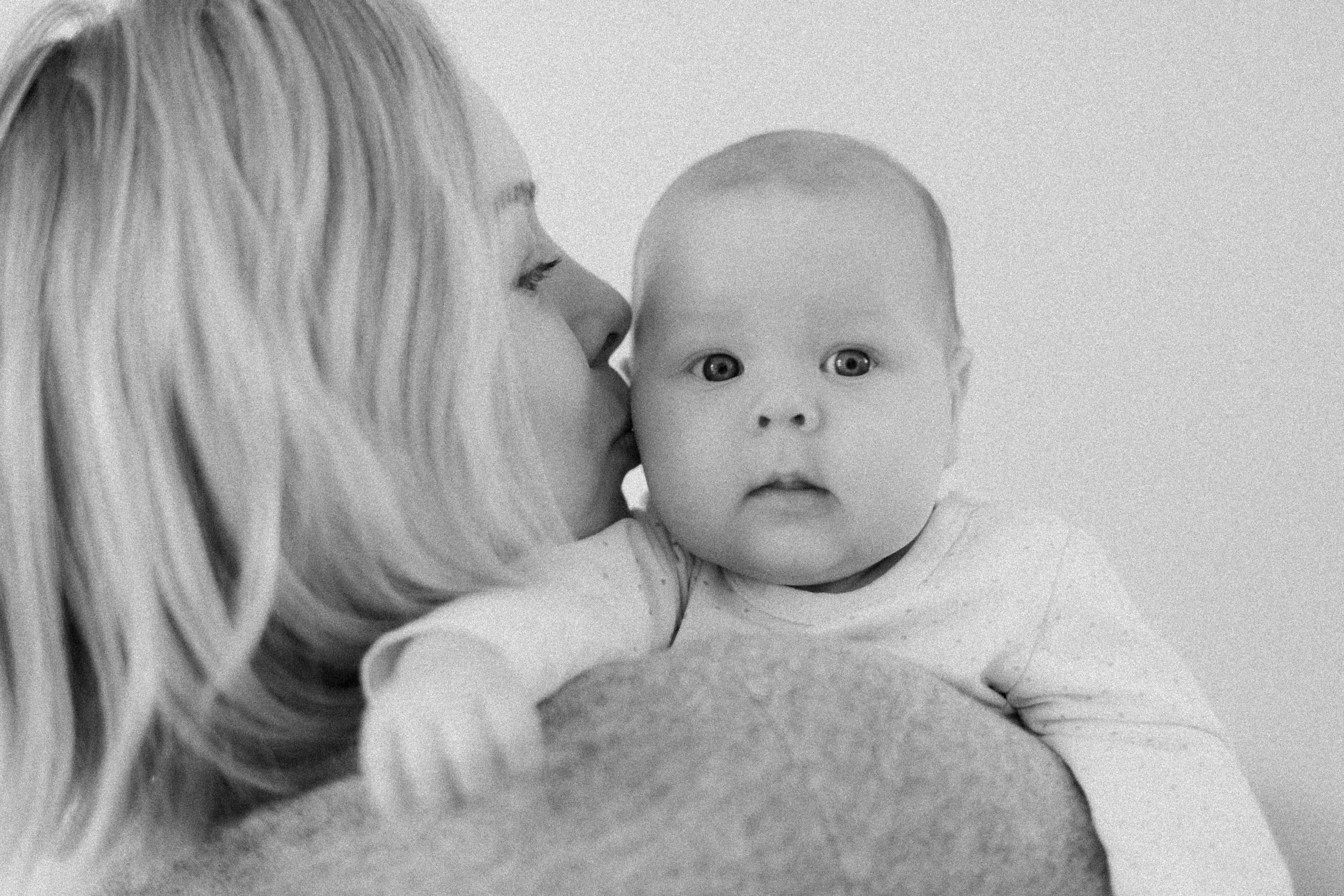 A woman with long blonde hair holding and kissing a baby with light skin and short hair, both in light-colored clothing, in a close-up black-and-white photograph.