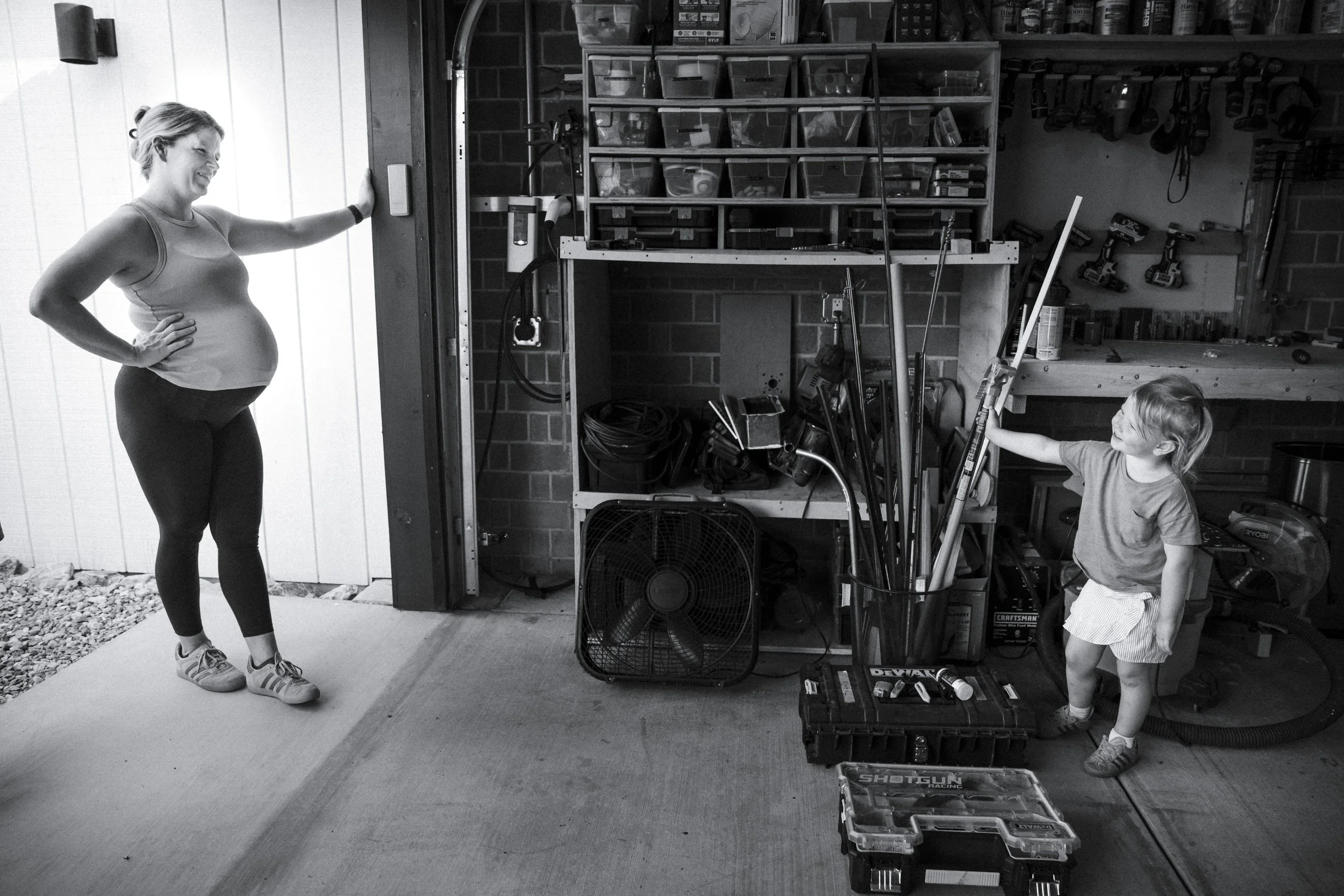 A woman and a young girl are in a garage, smiling and playing with toy swords. The woman is standing near the doorway, touching the wall, while the girl is holding a toy sword, pointing it toward the woman.