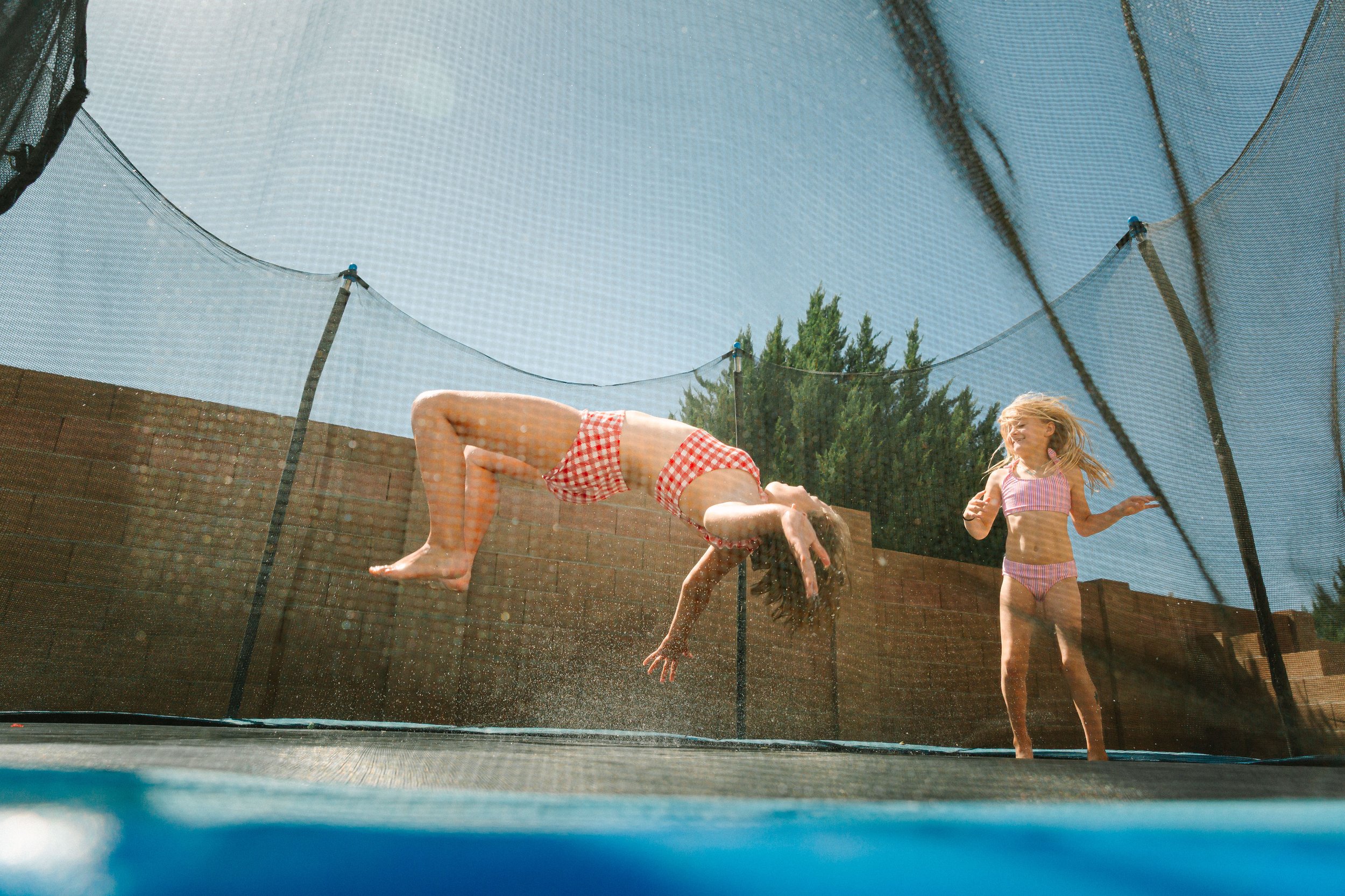 Two children, a boy and a girl, playing and jumping on a trampoline outside. The boy is upside down, and the girl is standing nearby smiling. They are wearing swimsuits, and there's a safety net around the trampoline with a brick wall and trees in the background.