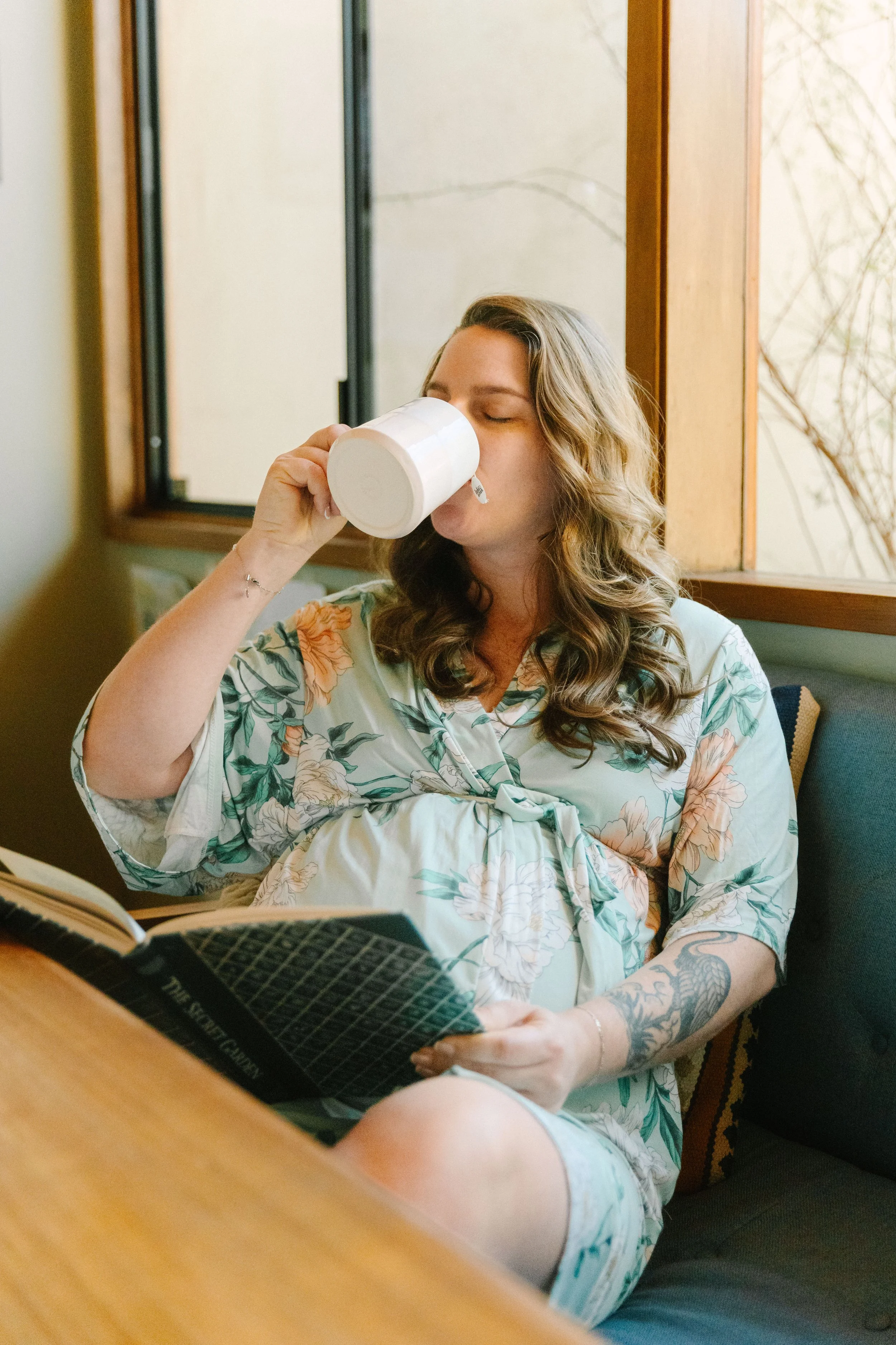 A woman with wavy hair and tattoos on her arm is sitting indoors by a window, drinking from a white mug while reading a book.
