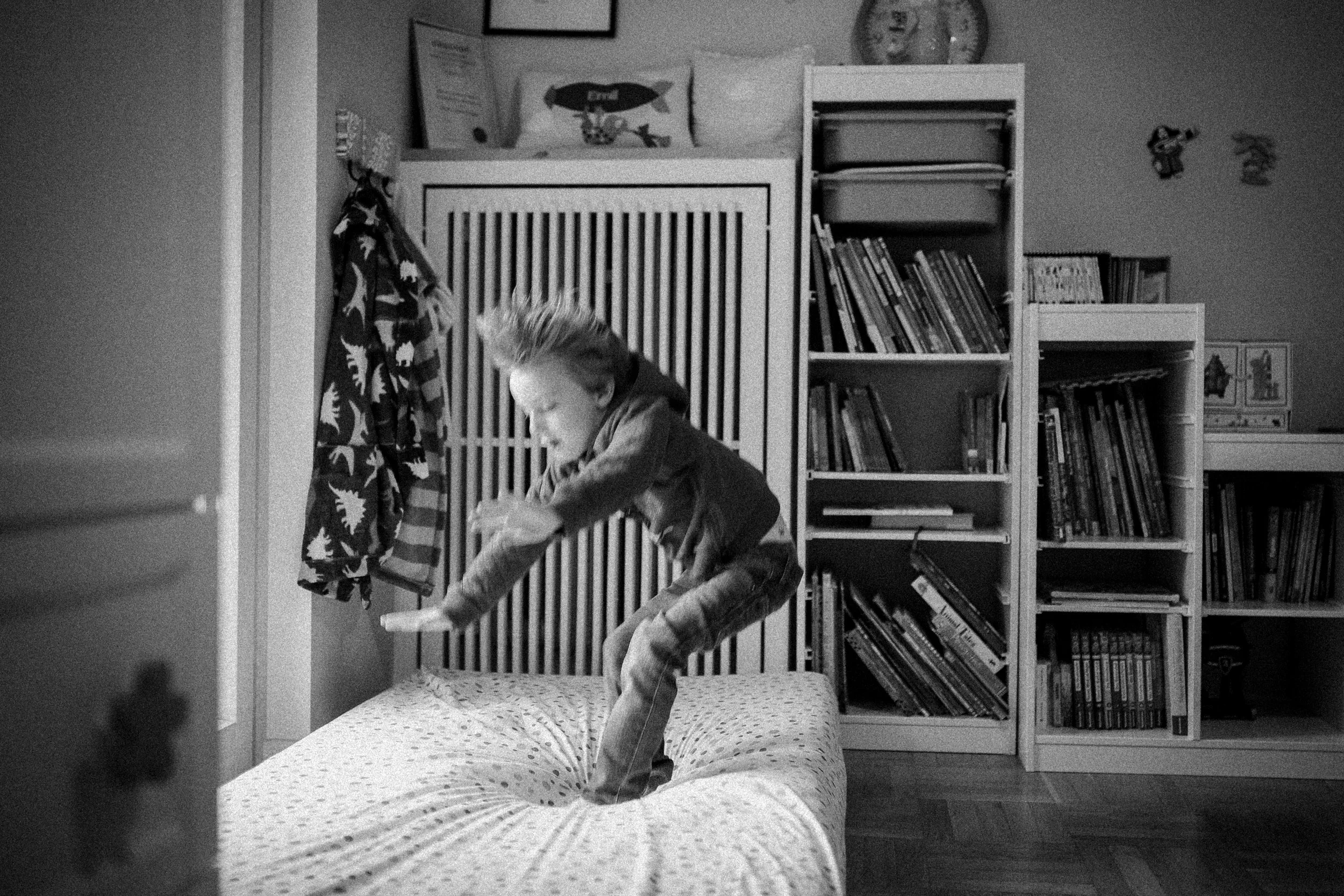 A young boy with light-colored hair jumping on a bed in a room with bookshelves, standing on a bed with a polka dot cover . The room has a clock on the wall, a framed certificate, and a blanket with a dinosaur pattern hanging nearby.