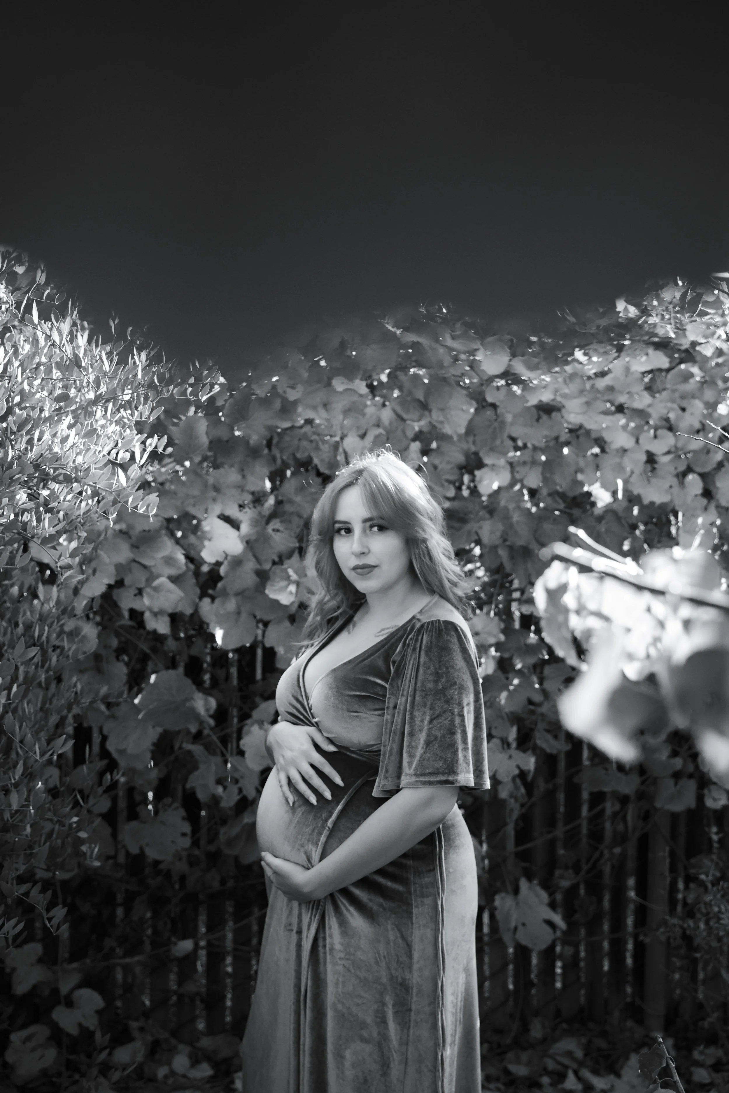 Black and white photo of a pregnant woman standing outdoors among leafy plants, gently cradling her baby bump with her left hand, with a confident expression on her face.