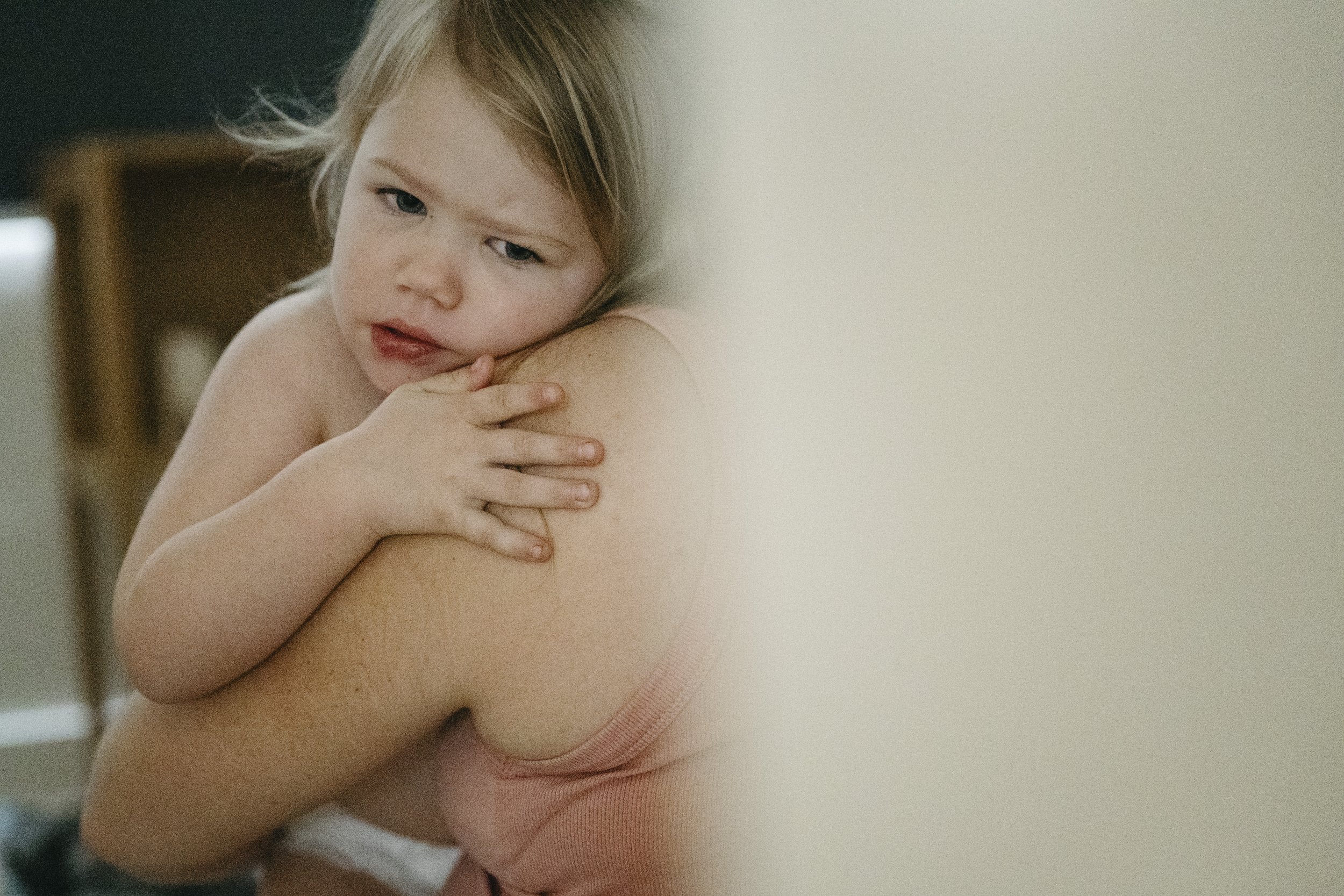 A young child with light brown hair resting their head on an adult's shoulder, with one hand touching the adult's arm. The child has a slightly confused or curious expression.