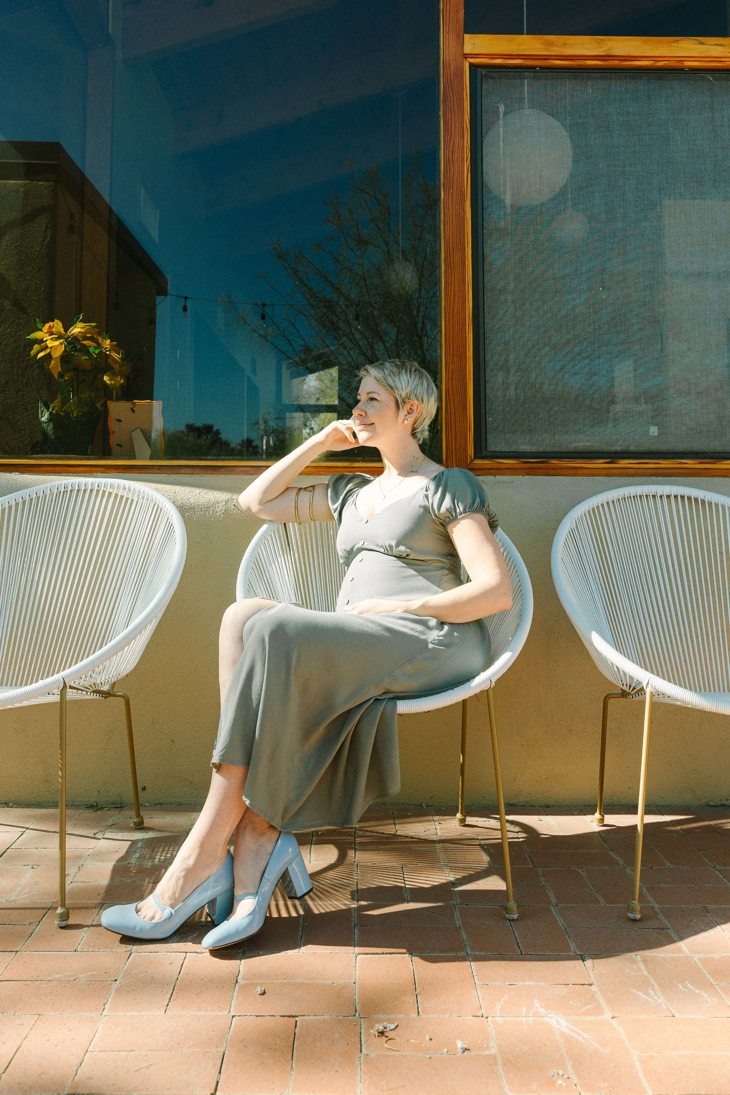 A woman with short gray hair and light skin, sitting on a white modern outdoor chair, wearing a gray dress and matching heels, smiling while enjoying sunlight on a balcony with a glass window and potted plant in the background.