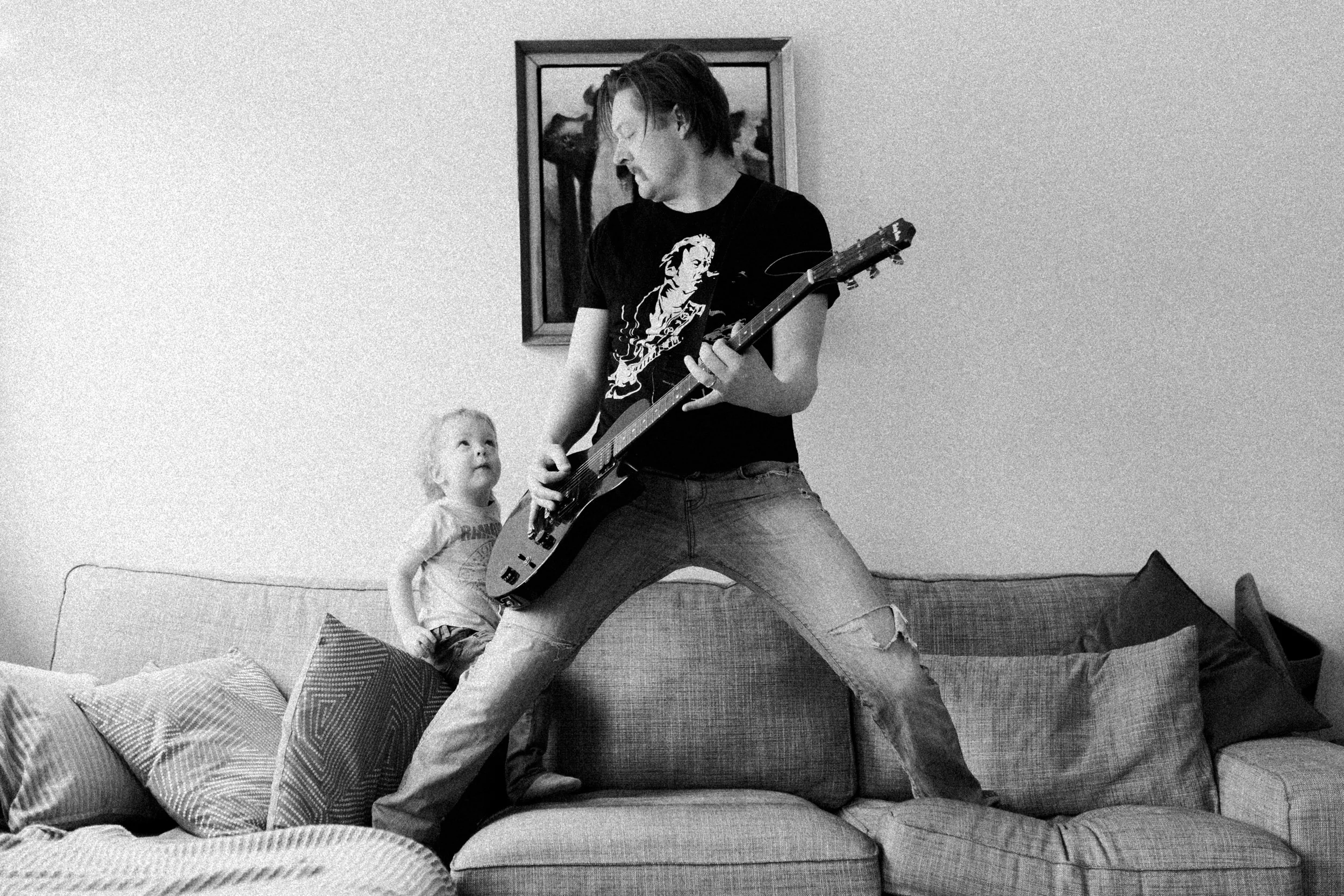 A man playing an electric guitar on a couch with a child watching beside him, in a living room with a framed picture on the wall.