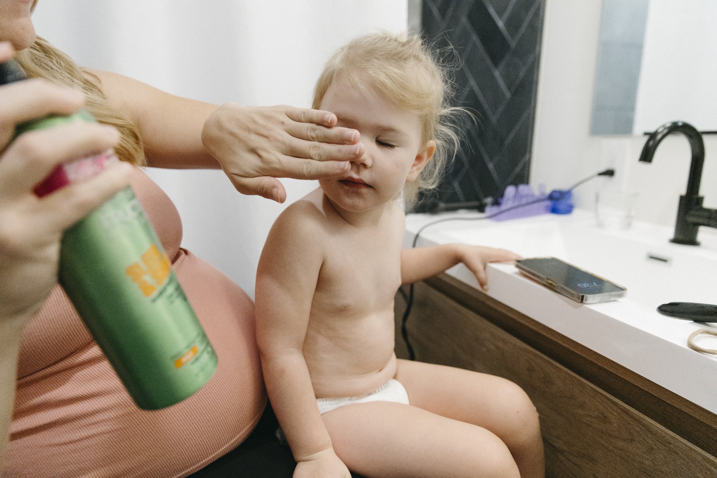 A young child sitting by a bathroom sink with an adult applying lotion or cream on their face. The child is shirtless and wearing a diaper, with a phone and some toiletries on the sink.