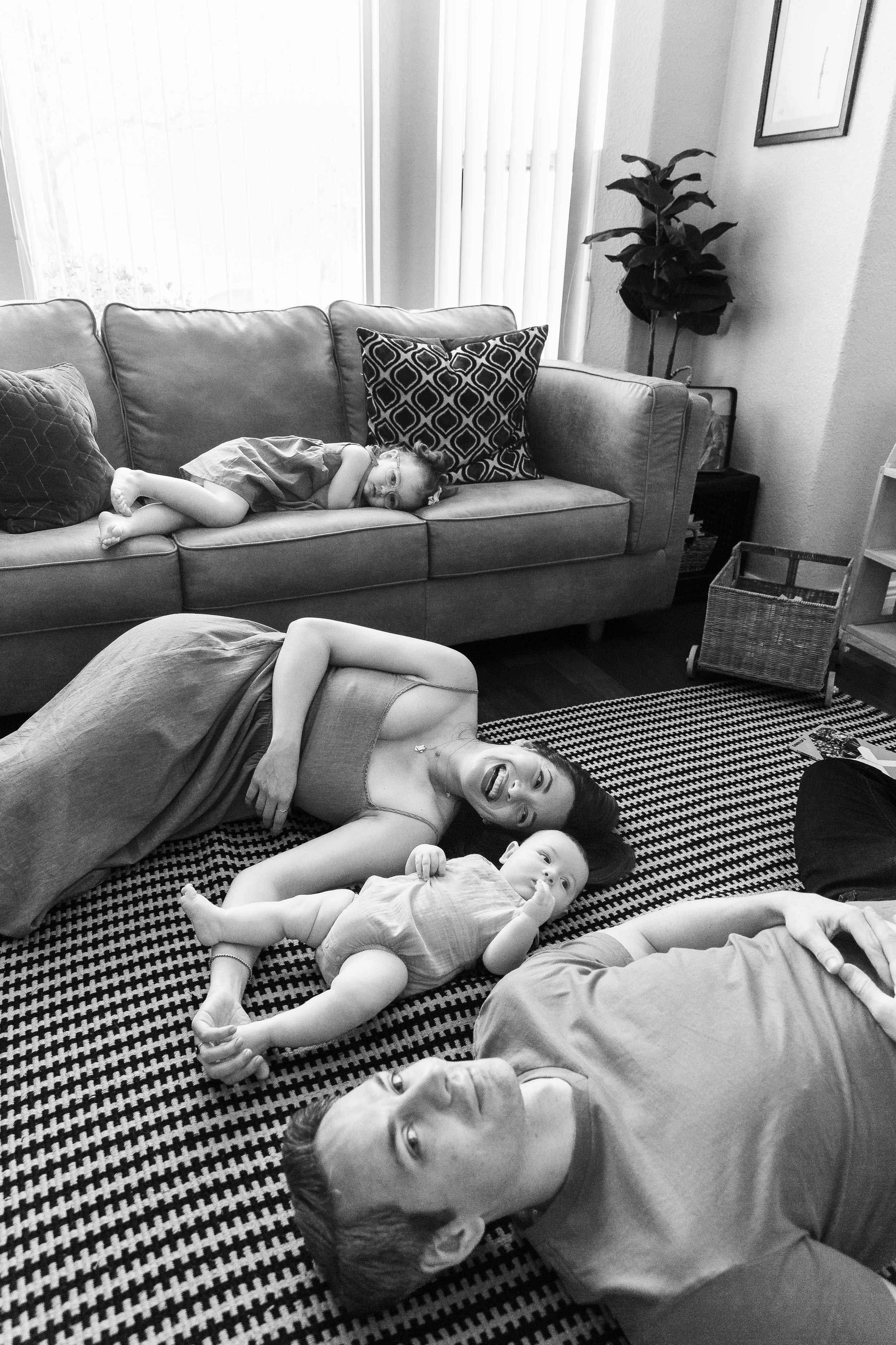 Black and white photo of multiple children lying on a patterned rug, some on a sofa, in a living room with a window, curtains, a wall with framed pictures, a side table, and a plant.