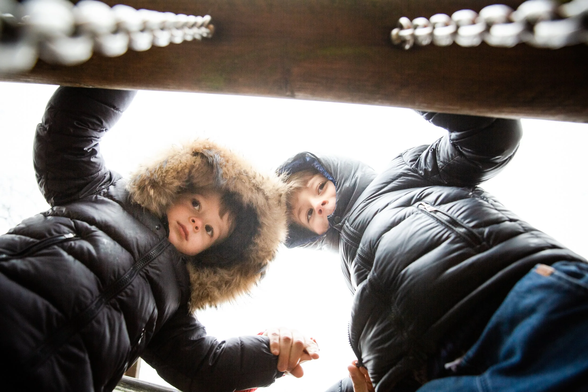 Two children wearing winter jackets with hoods, one with fur trim, looking down into the camera that is positioned below them, outdoors with overcast sky.