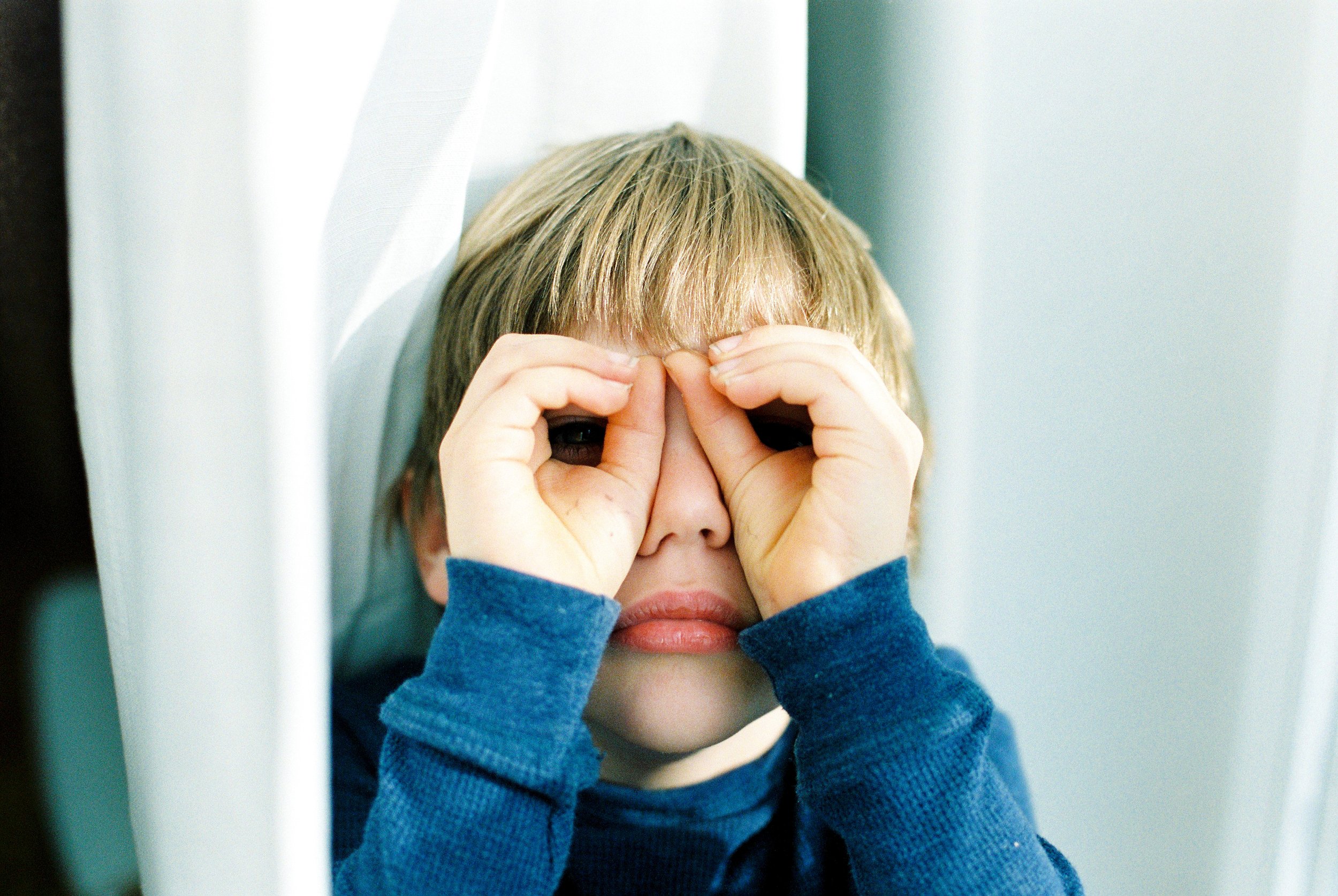 A young boy with blonde hair wearing a blue long-sleeve shirt making binoculars with his hands around his eyes.