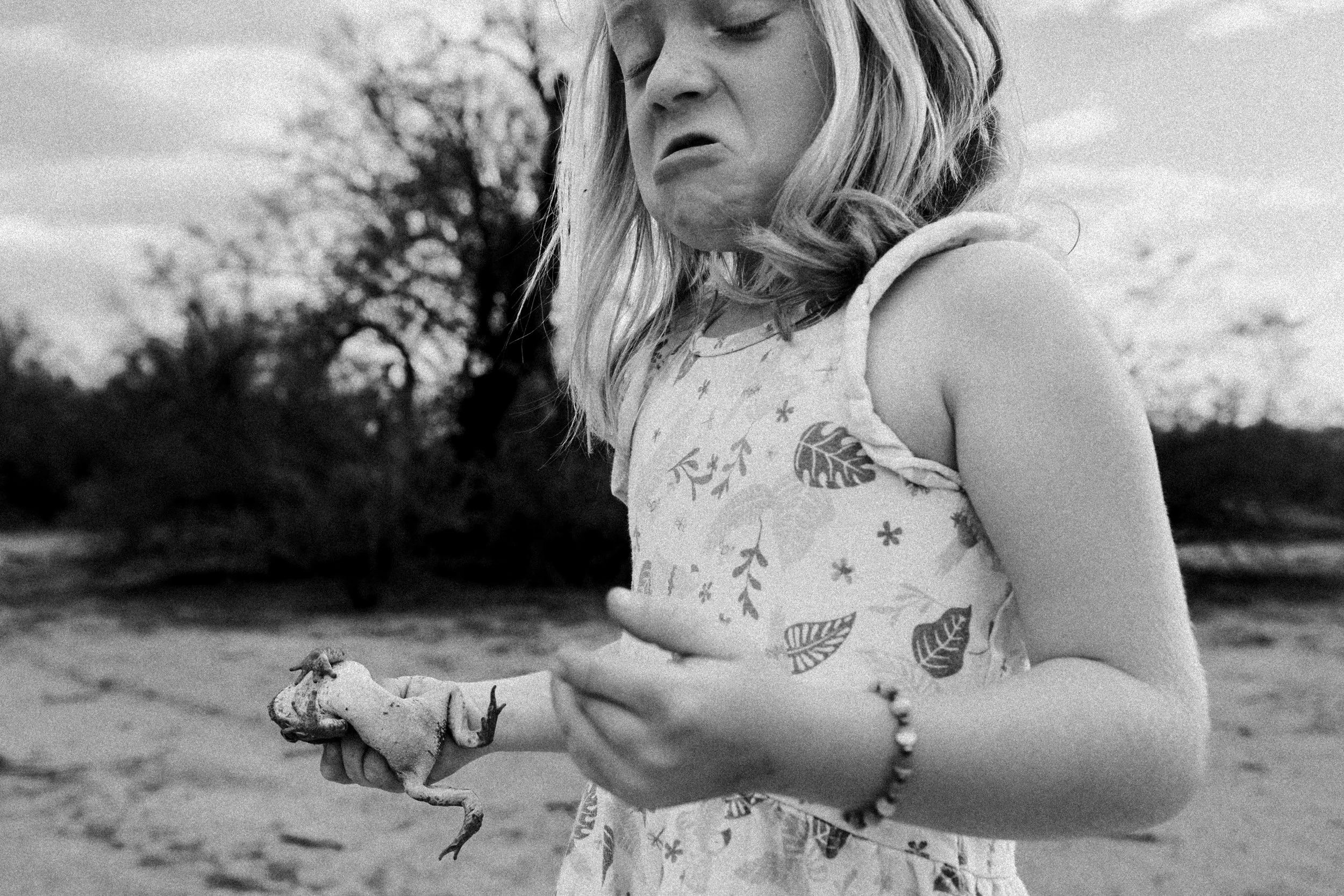 A young girl with long, wavy hair is outdoors, holding a toad. She is wearing a short-sleeved dress with leaf patterns and appears to be disgusted by the toad.  The background shows trees and an open sky.