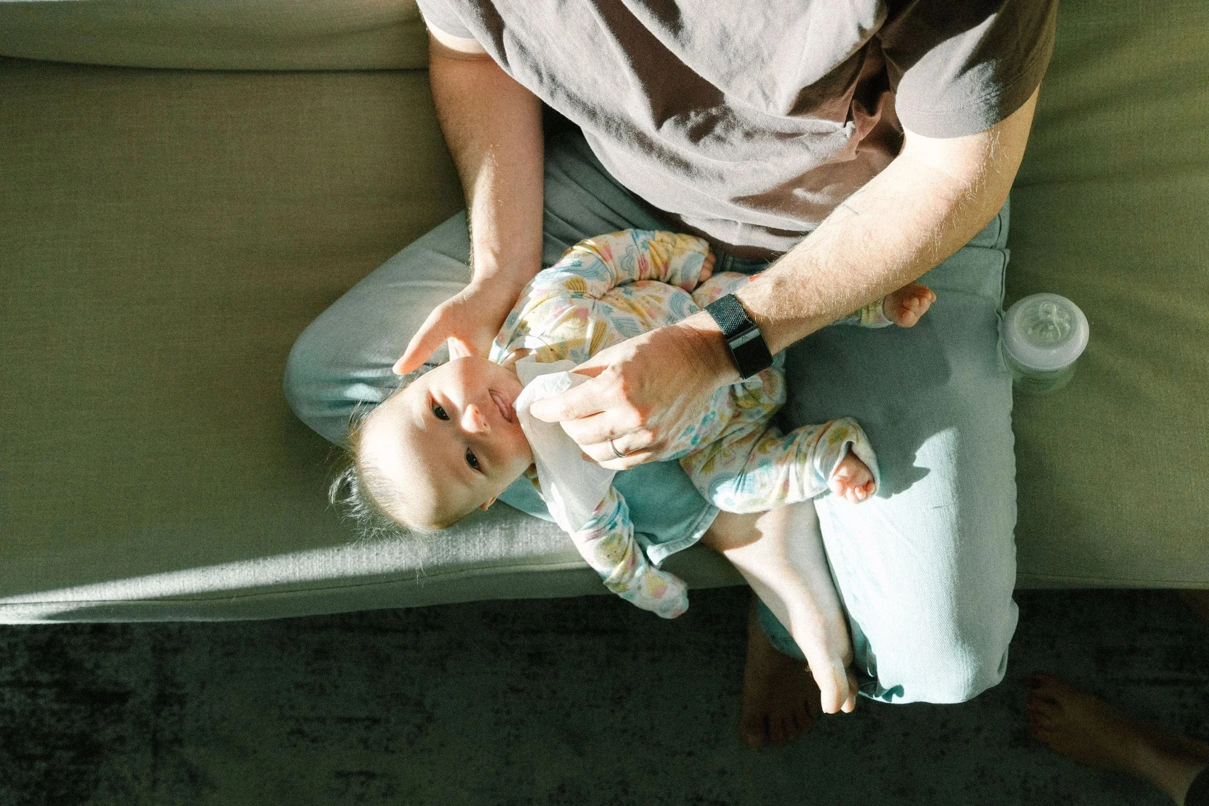Child laying on a couch with a man, possibly a father, holding a tissue or cloth to the child's face. The man is wearing a watch and a casual t-shirt. There is a water bottle on the couch near them, and the child is wearing a colorful patterned pajama top.