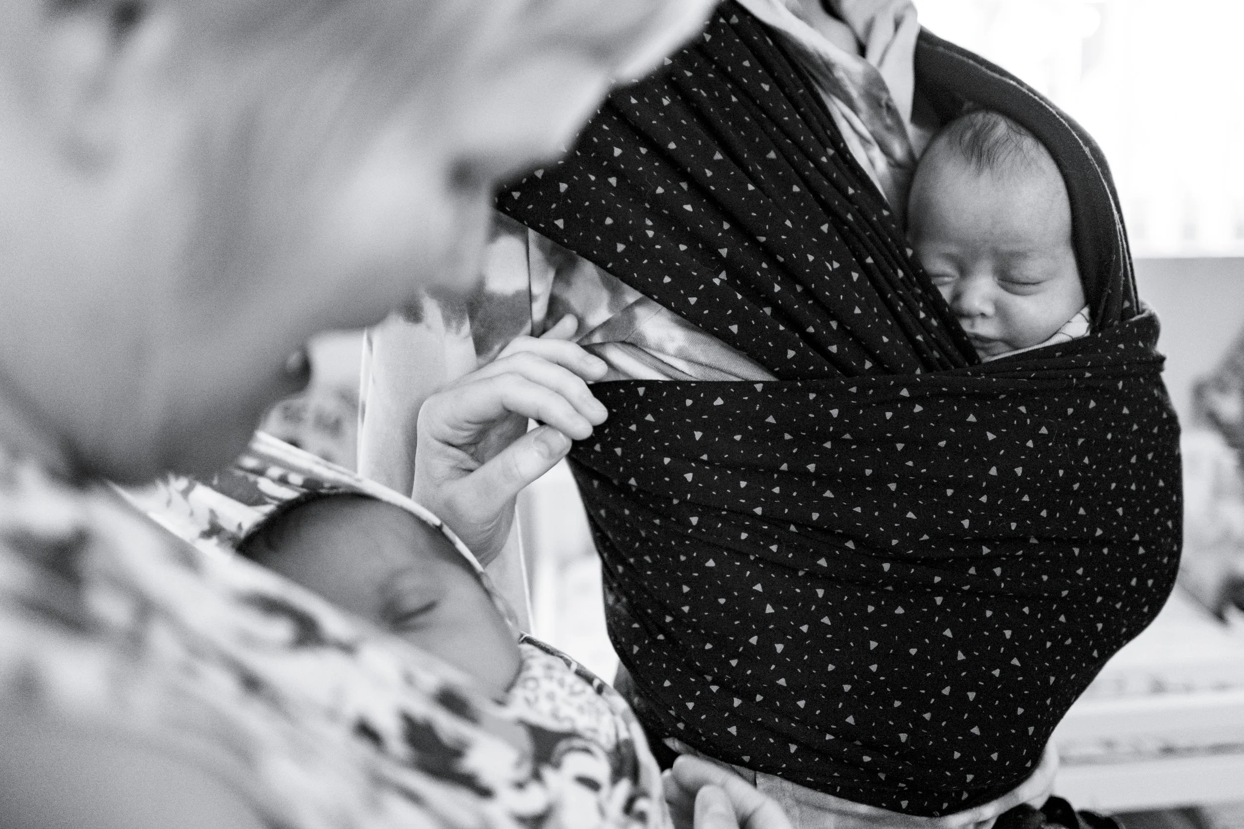 A woman holding a sleeping baby wrapped in a patterned wrap carrier, with part of a child's face and hand visible in the foreground.