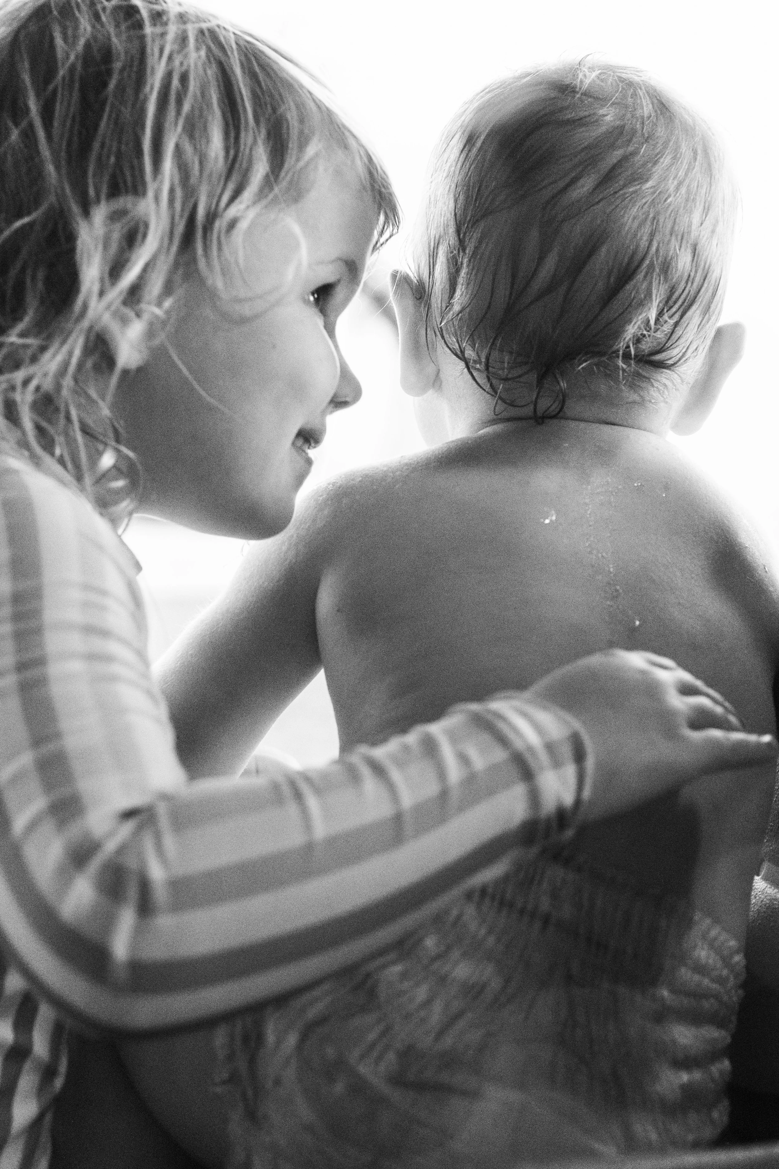 A woman with curly hair and a child with wet hair are close together, facing each other, with the woman smiling as she looks at the child. The child is shirtless and appears to be holding onto or leaning on an object with a checkered pattern.