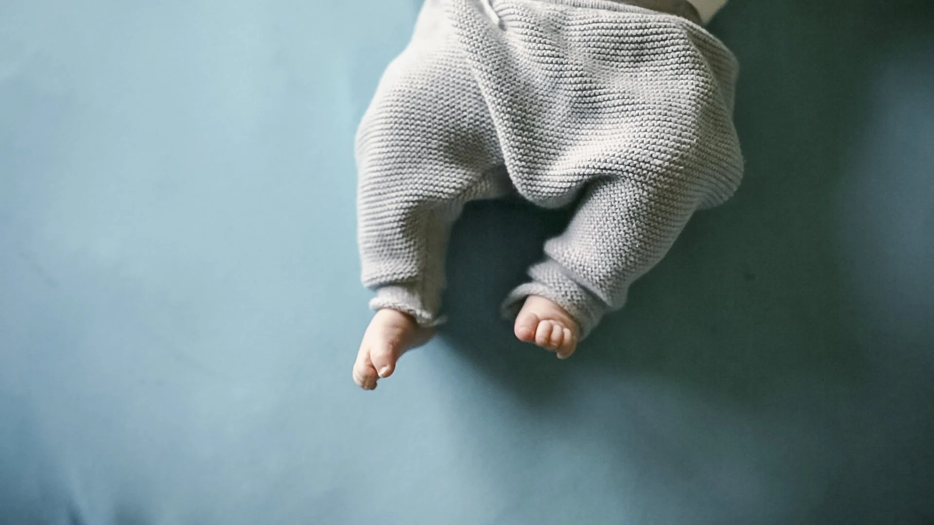 Close-up of a baby crawling on a blue surface, wearing a long-sleeved shirt and dark pants, with their hand reaching forward.