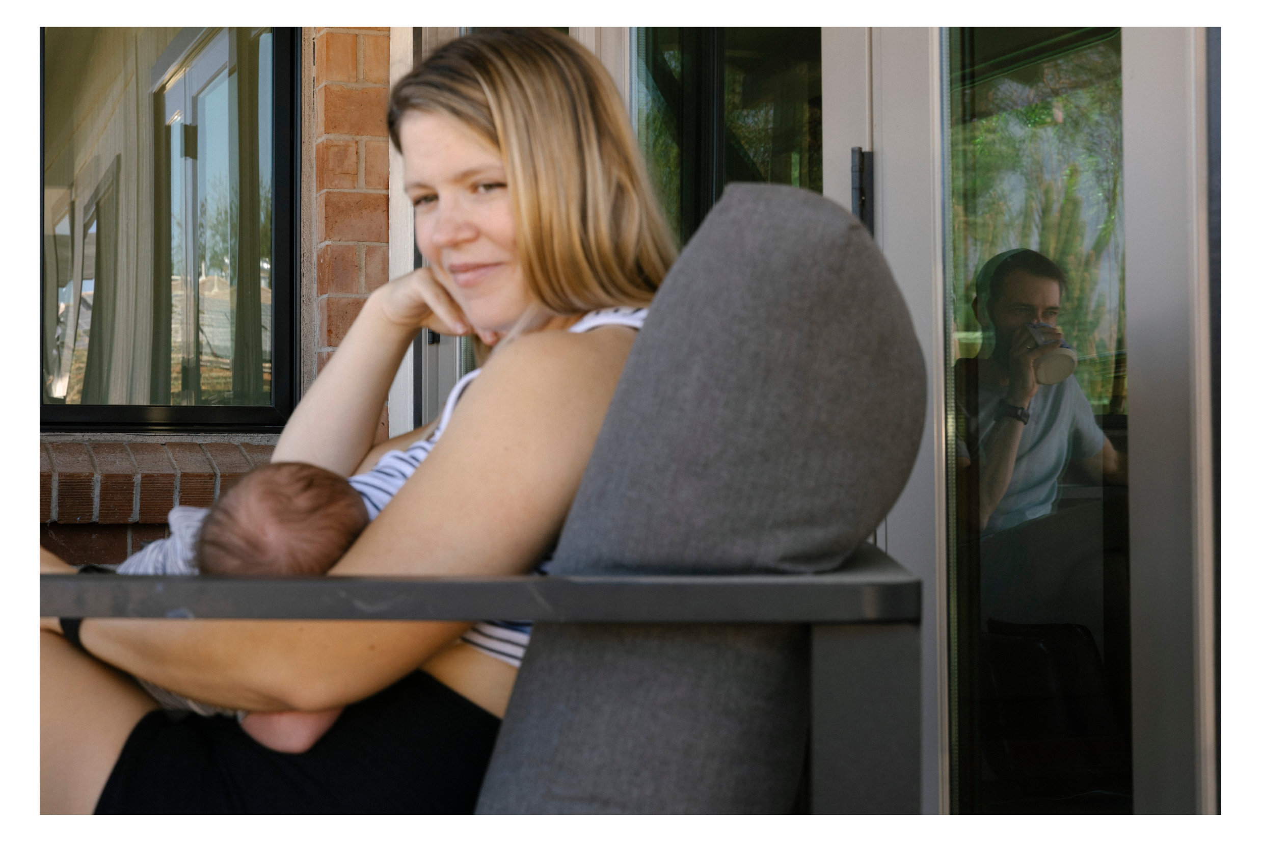 mom nursing her newborn while watching her toddler play in the yard and dad is in the reflection drinking coffee at in home newborn session downtown tucson