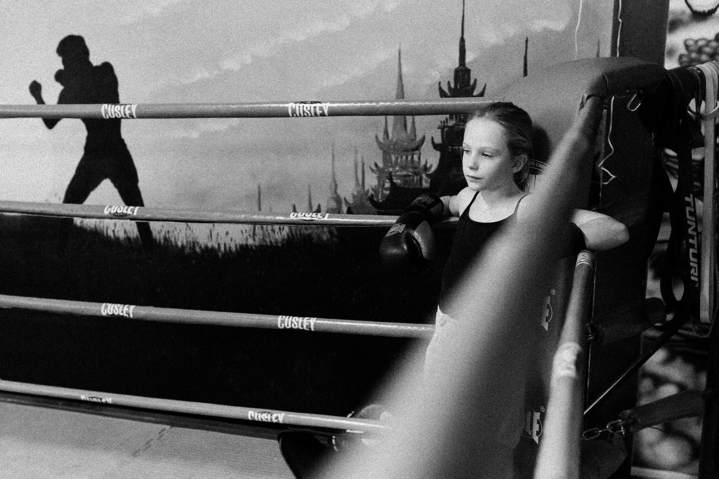 A young girl with boxing gloves resting on a boxing ring corner post, looking thoughtful, with a silhouette of a person walking in the background and a castle silhouette in the sky.