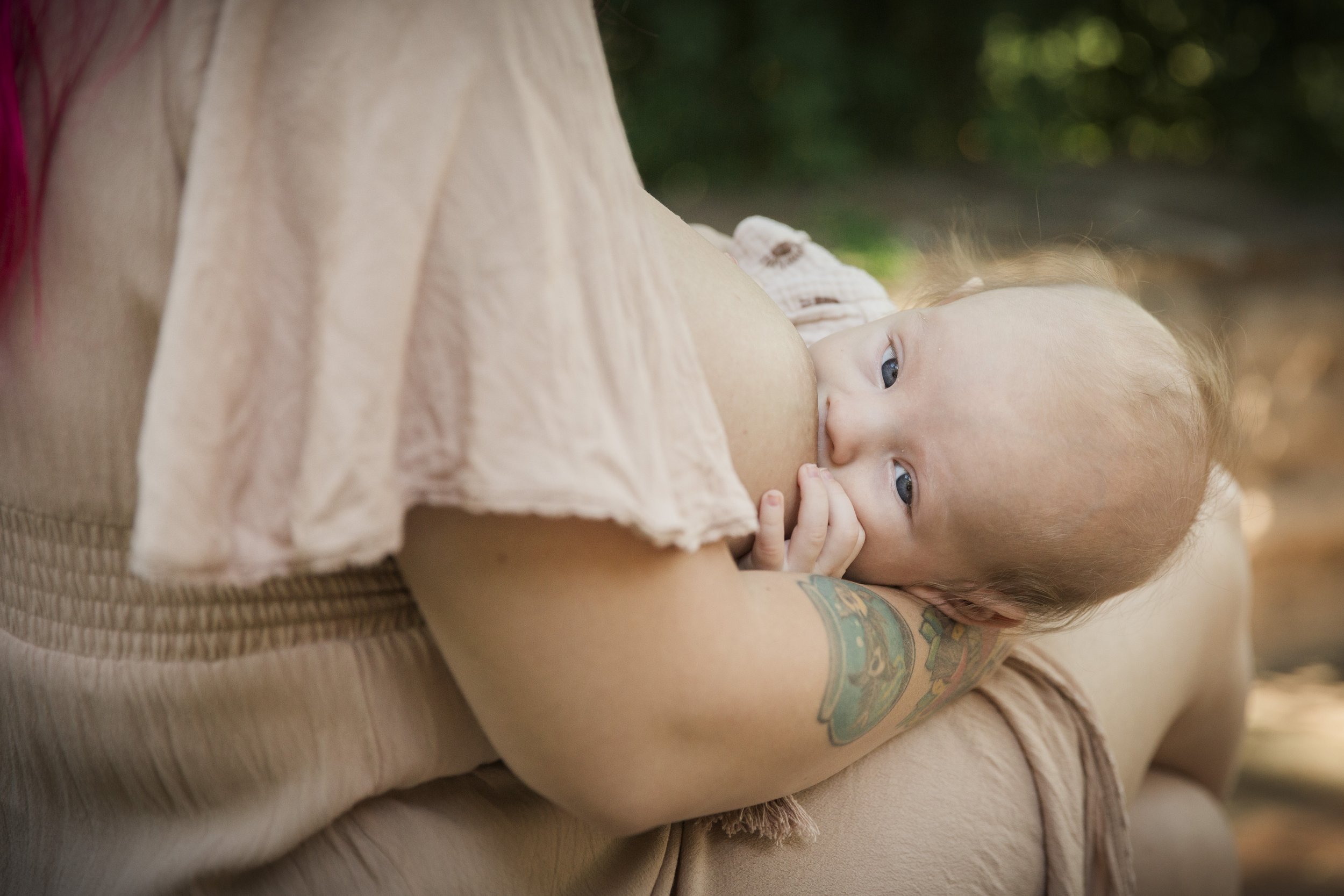 A baby girl with blue eyes nursing from her mother outdoors with greenery in the background.