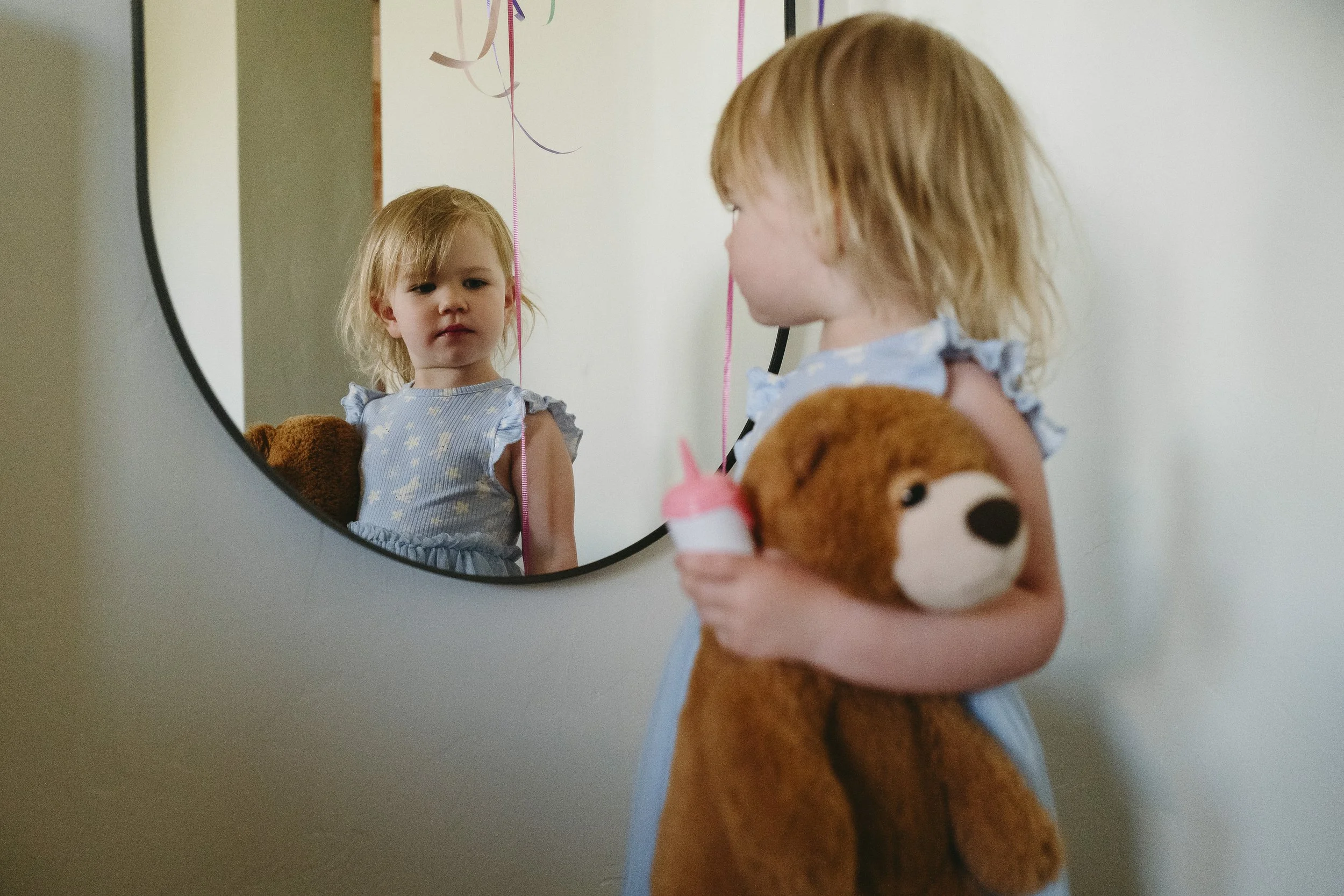 A young girl looking at herself in a round mirror, holding a teddy bear and a small bottle, with colorful ribbons hanging on the mirror