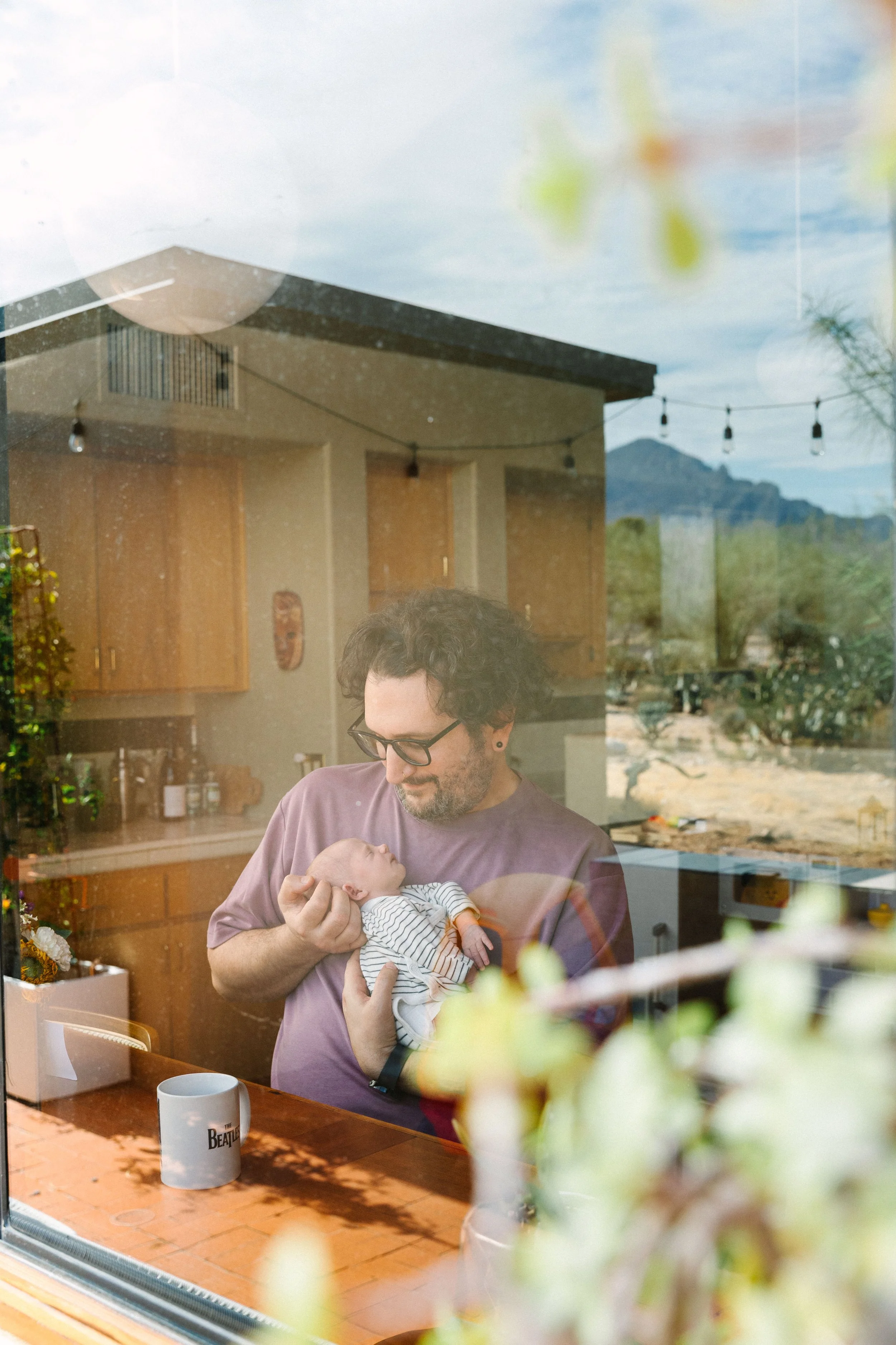 A man with black curly hair, glasses, and earrings is holding a baby with a striped onesie inside a room, viewed through a window with reflections of the outdoor scenery including desert landscape, mountains, and string lights.