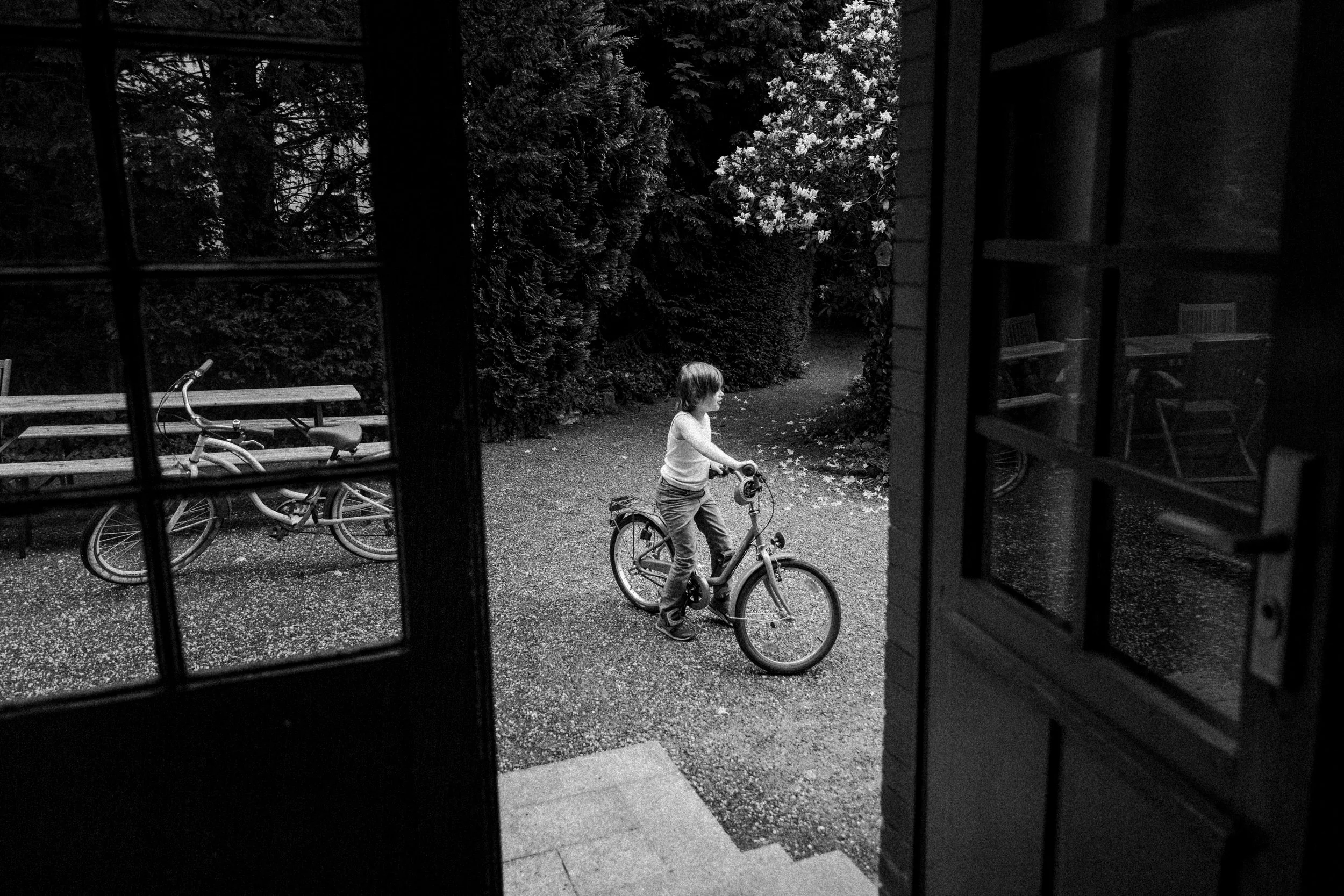 View through a doorway showing a young boy riding a bicycle outdoors, with bicycles and greenery visible in the background.