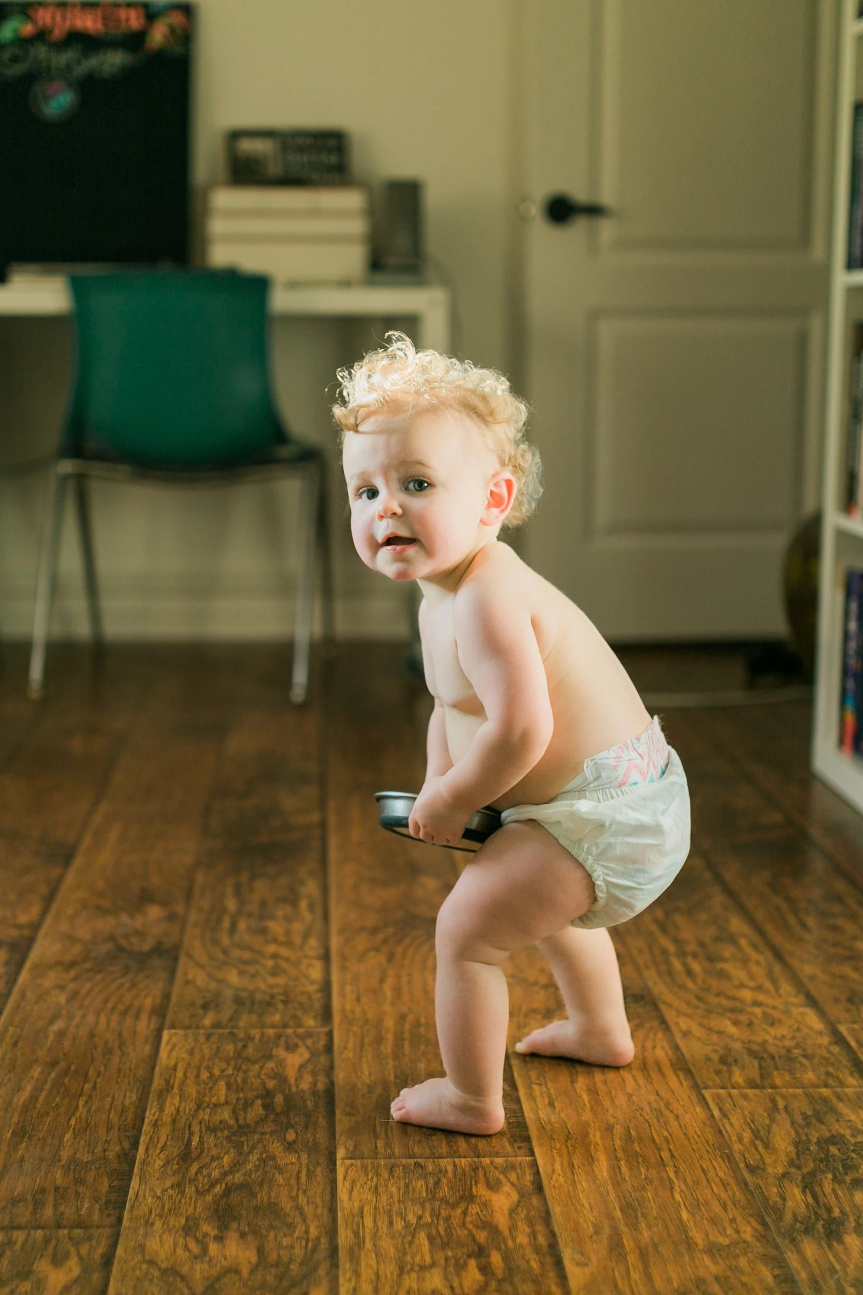 A young child with curly blonde hair, wearing a diaper, standing on a wooden floor, holding onto an object, with a blurred background of a room with chairs, books, and a door.