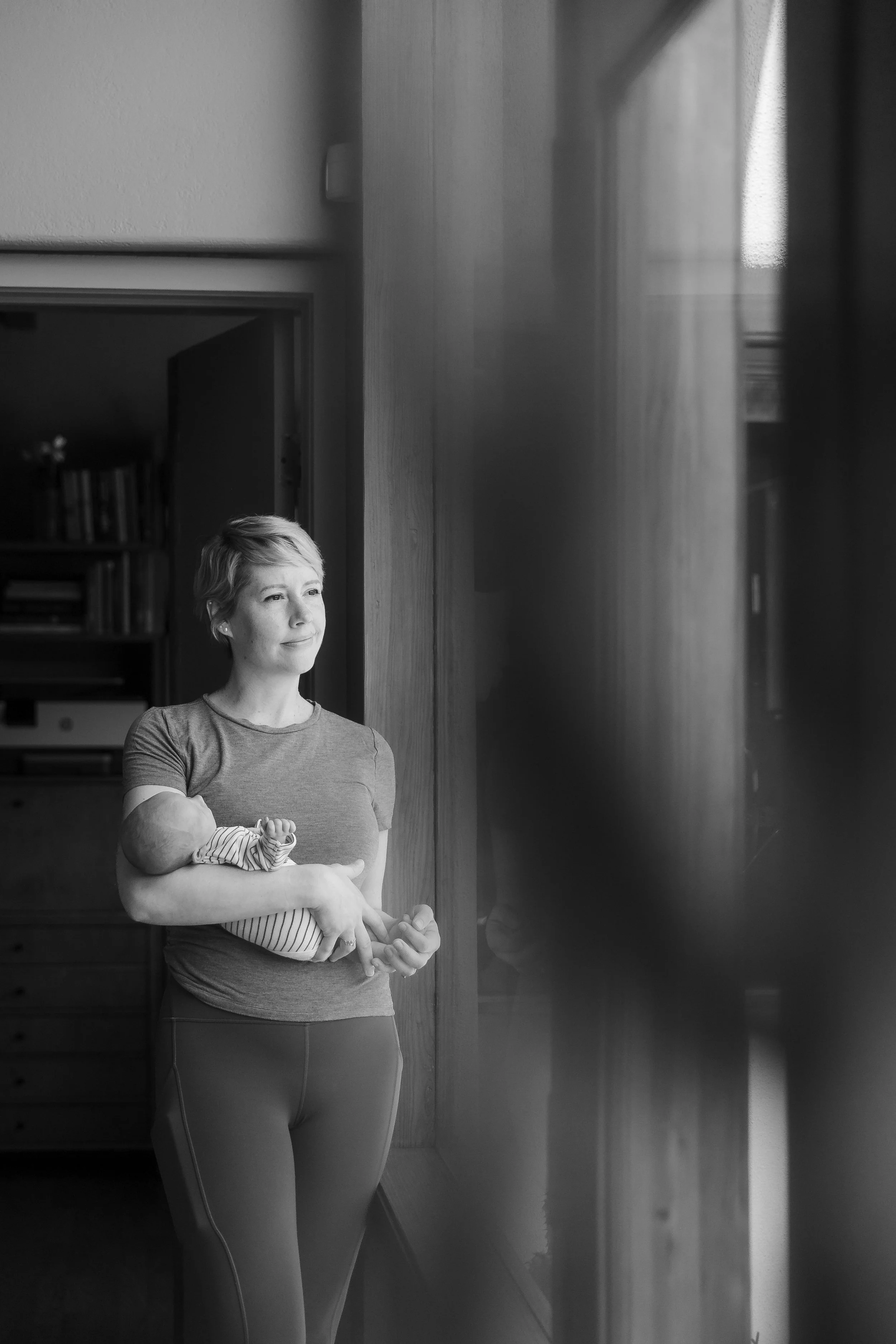 A woman with blonde hair holding a baby, standing indoors near a wooden wall and a glass window, in black and white.