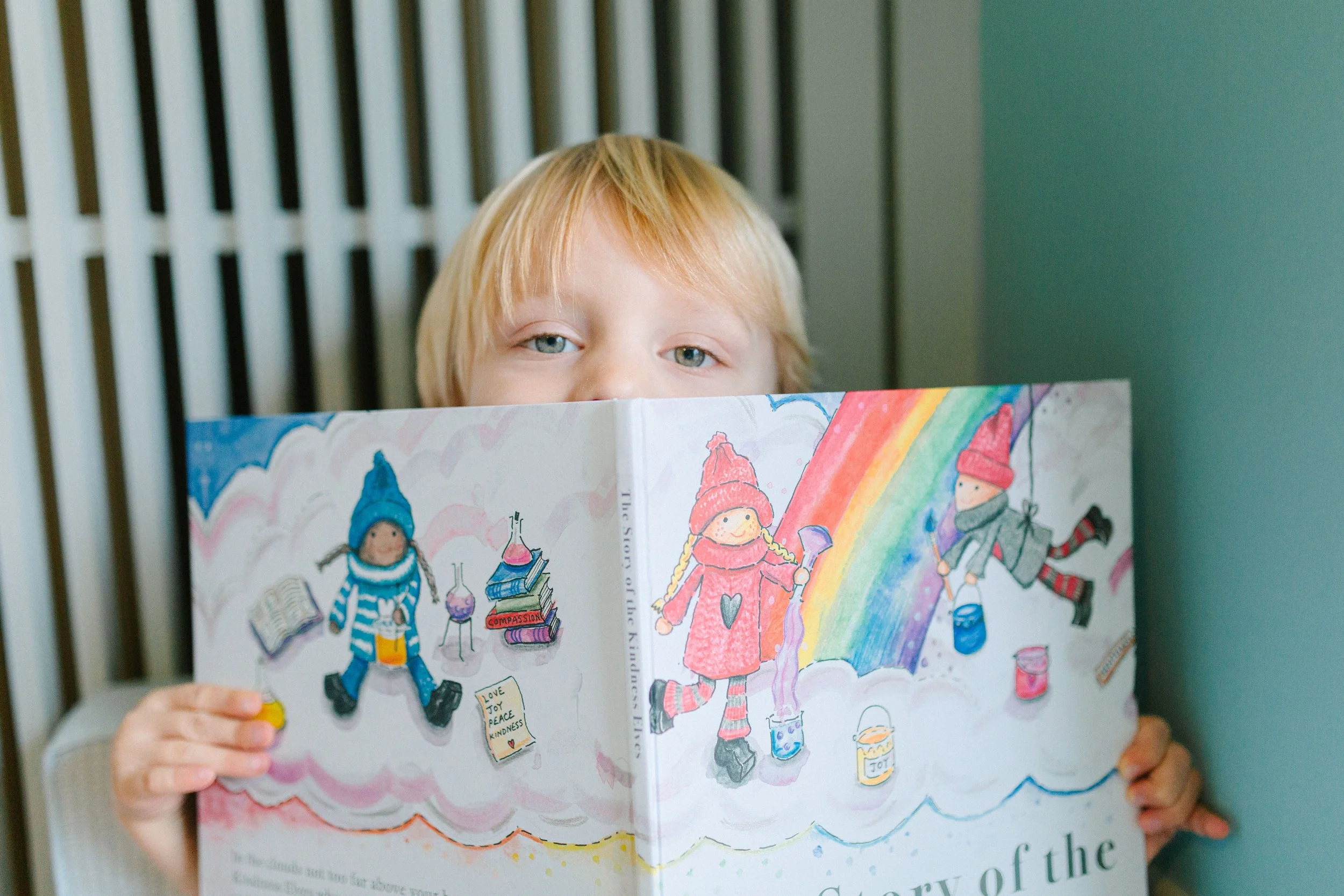 A young child with blonde hair and blue eyes is holding a colorful drawing or coloring book in front of their face, partially covering it. The drawing features children in winter clothing playing with paint and other art supplies, with a rainbow in the background.