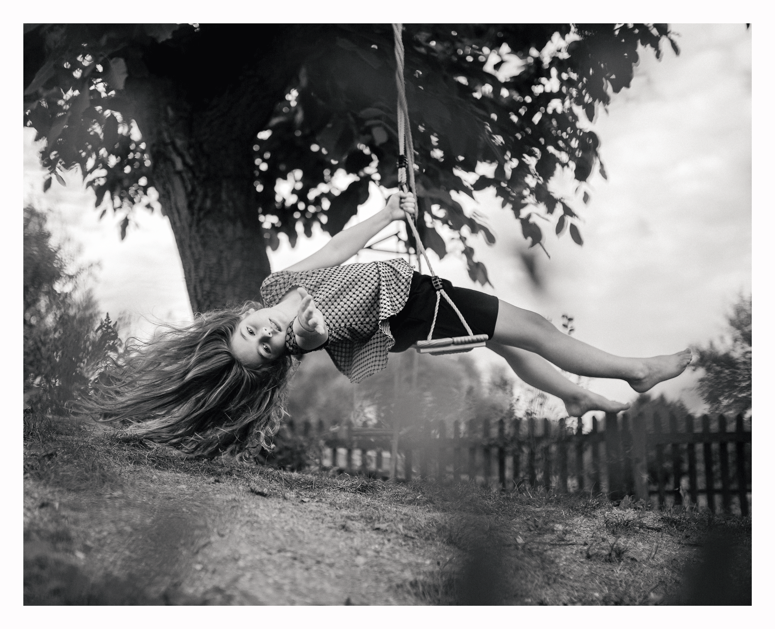 A young girl with long hair lying on the grass in front of a tree, pointing up, in black and white photography.