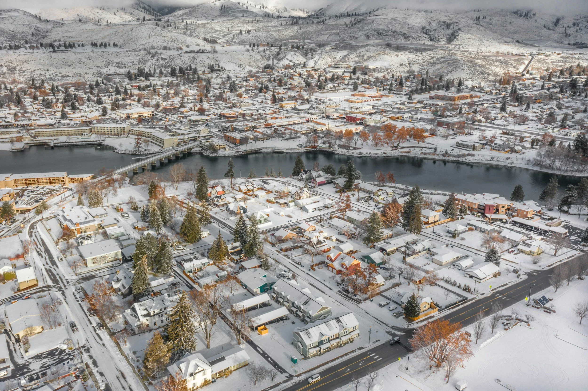 Aerial view of a snow-covered downtown Chelan WA. with houses, streets, and a river running through it, surrounded by the snow-covered Cascade mountains.