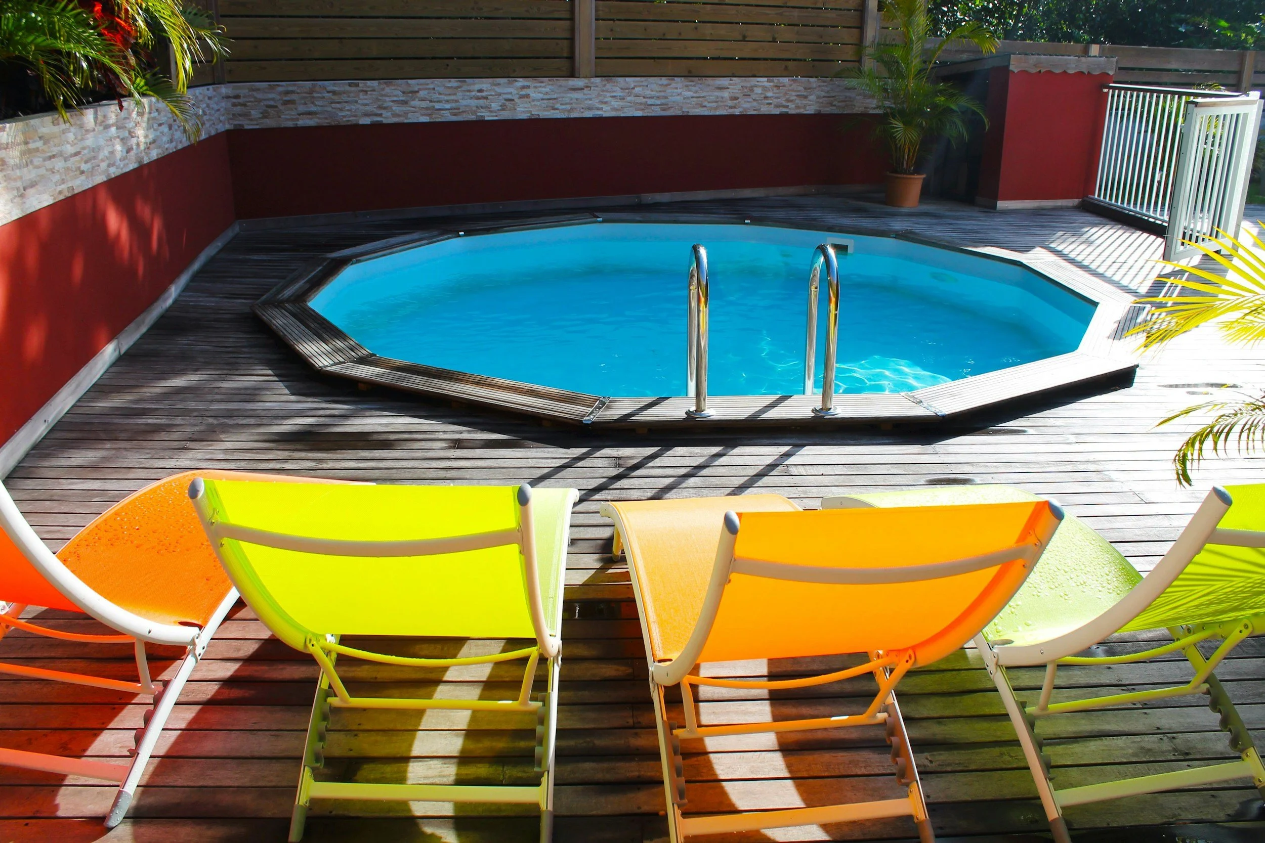 A small octagonal swimming pool with metal handrails, surrounded by a wooden deck with colorful lounge chairs and potted plants, enclosed by a wooden privacy fence.