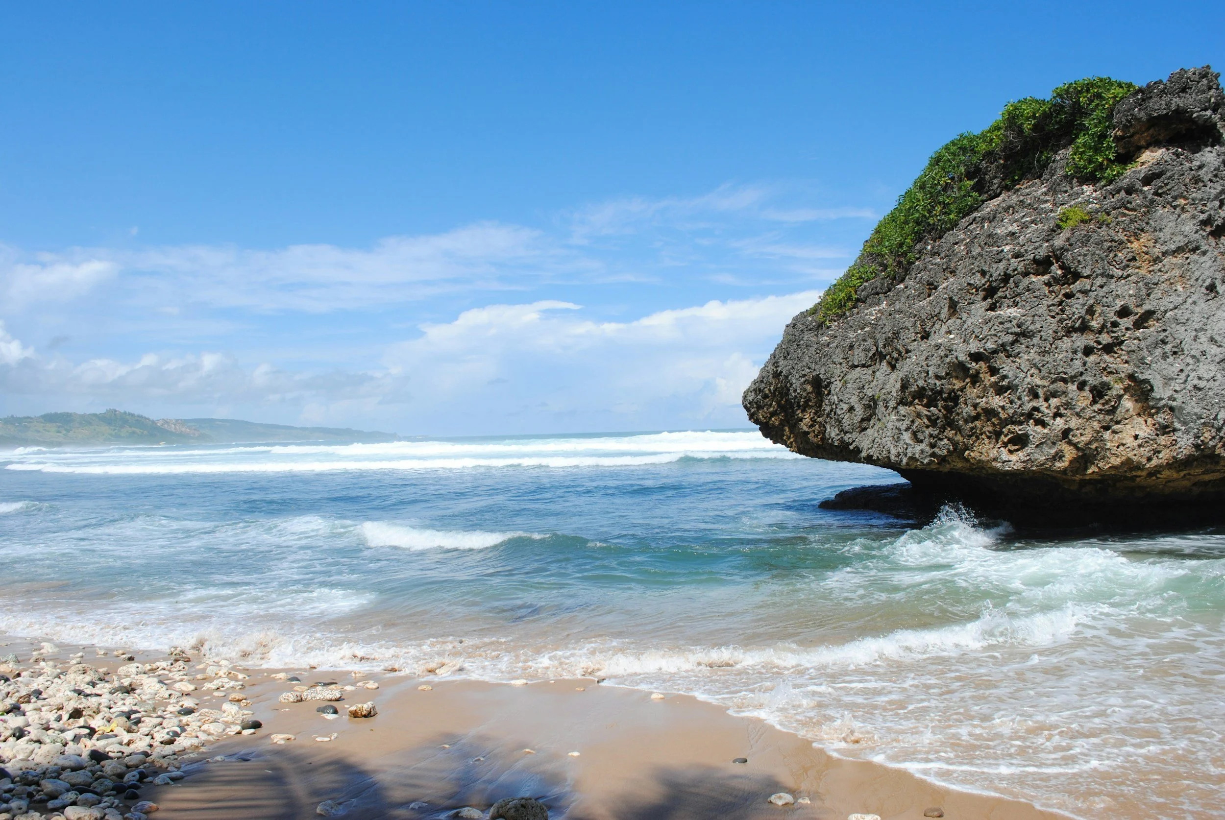 A tropical beach with a large rocky formation covered in green plants on the right side, sandy shoreline with pebbles, and clear blue ocean waves under a partly cloudy sky.