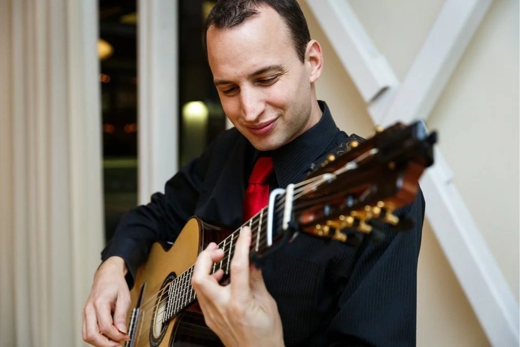 Man playing an acoustic guitar, wearing a black shirt and red tie, indoors near a window.