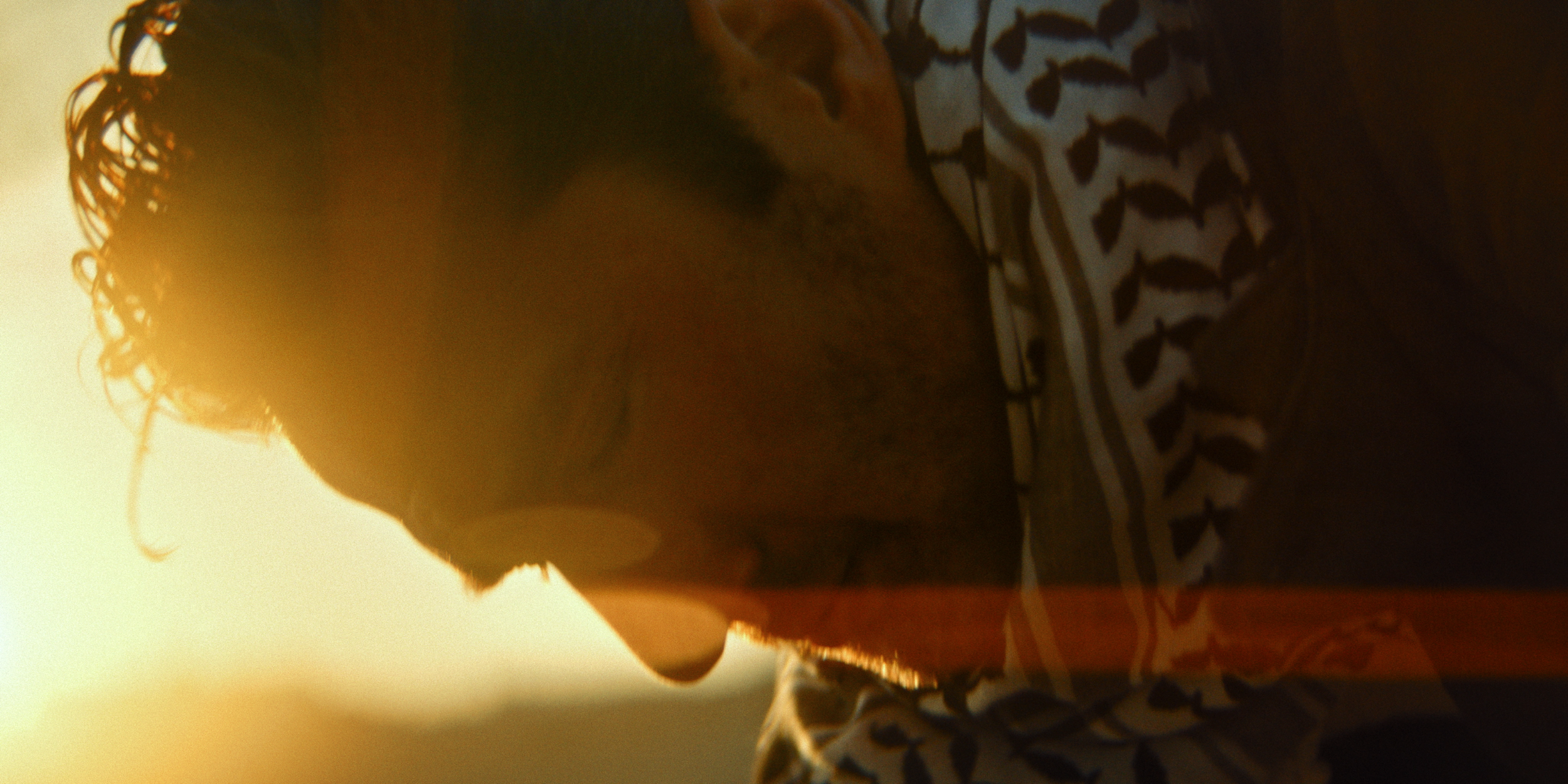 Close-up side profile of a man with curly hair and glasses, wearing a patterned shirt, in a warm backlit environment.