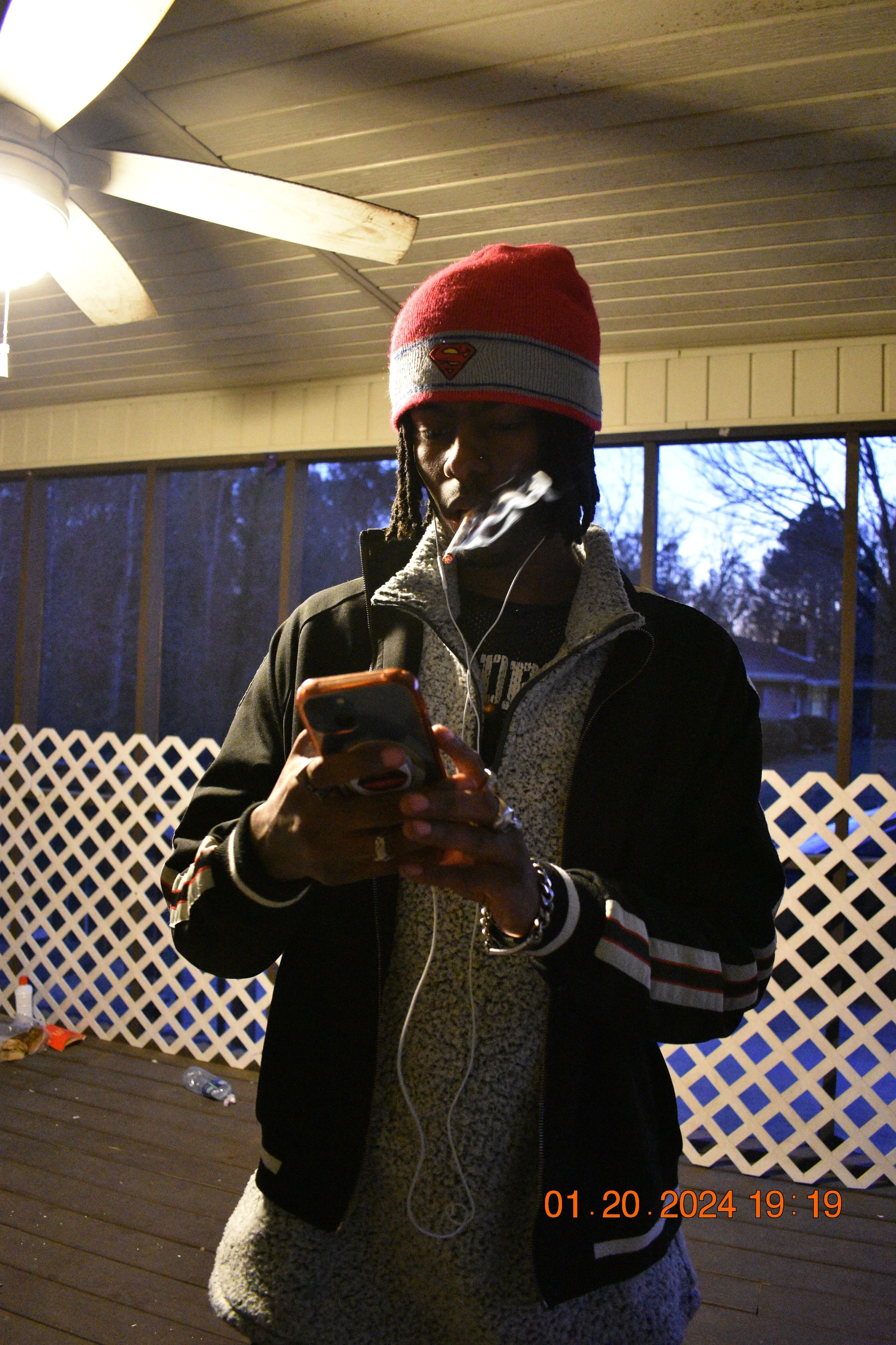 Young man with dreadlocks wearing a red Beanie with Superman logo, using smartphone outdoors at dusk, smoking, on a porch with lattice fence and ceiling fan.
