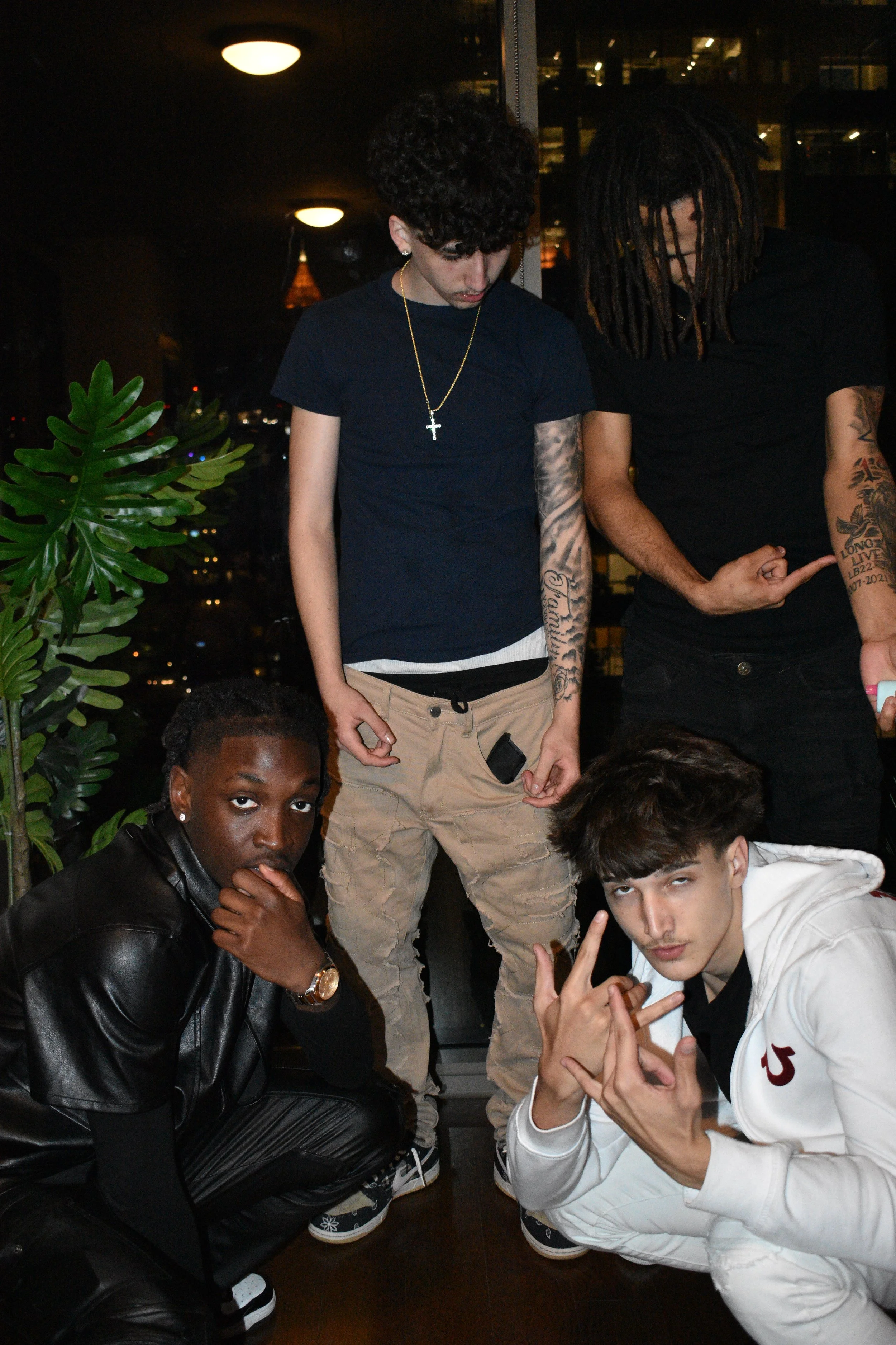 Four young men posing indoors at night with city lights in the background; one crouching, one kneeling, and two standing, all dressed casually.