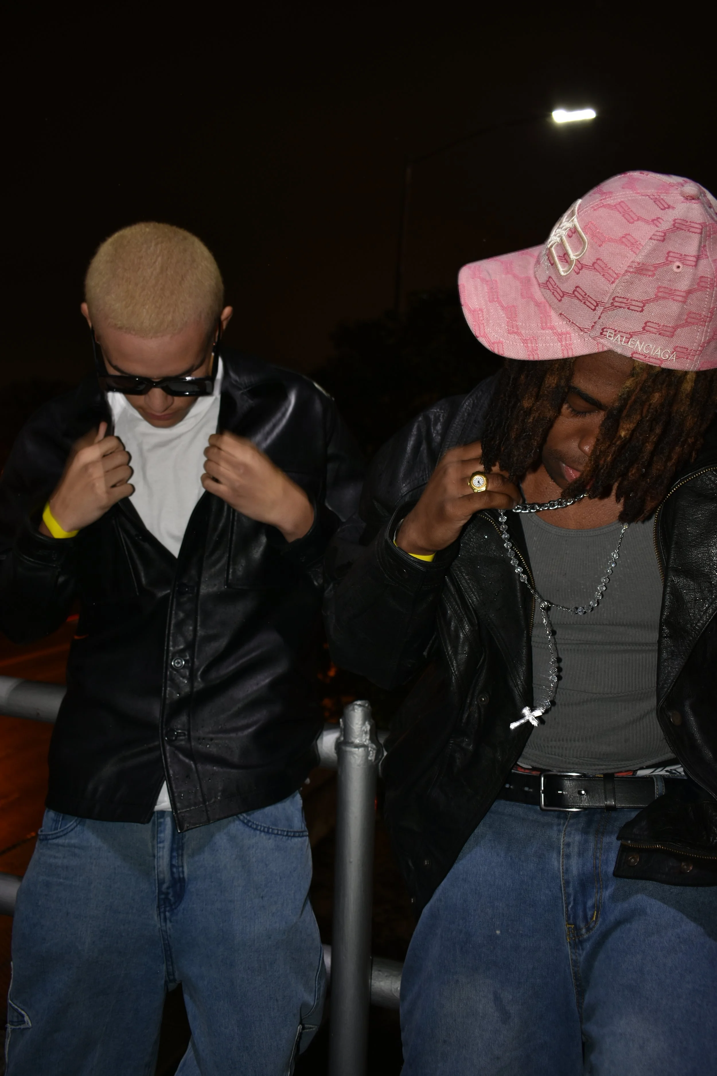 Two people standing outdoors at night, both wearing black leather jackets and blue jeans, with one wearing a pink cap and the other with dyed blonde hair and sunglasses.