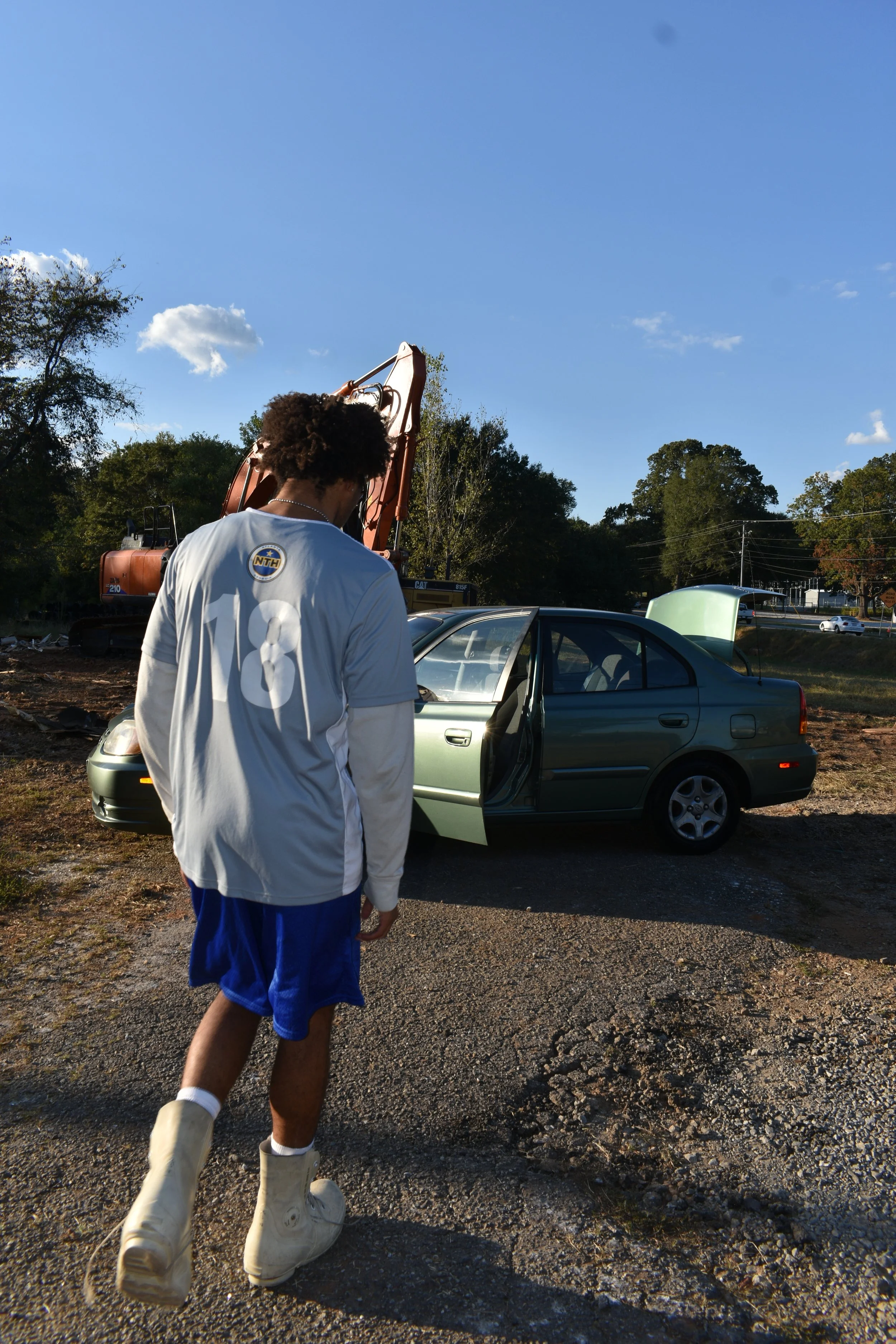 A person with curly hair wearing a blue sports jersey, blue shorts, and white boots walking away from a green car with its door open and hood raised, in a dirt lot with construction equipment and trees in the background on a sunny day.