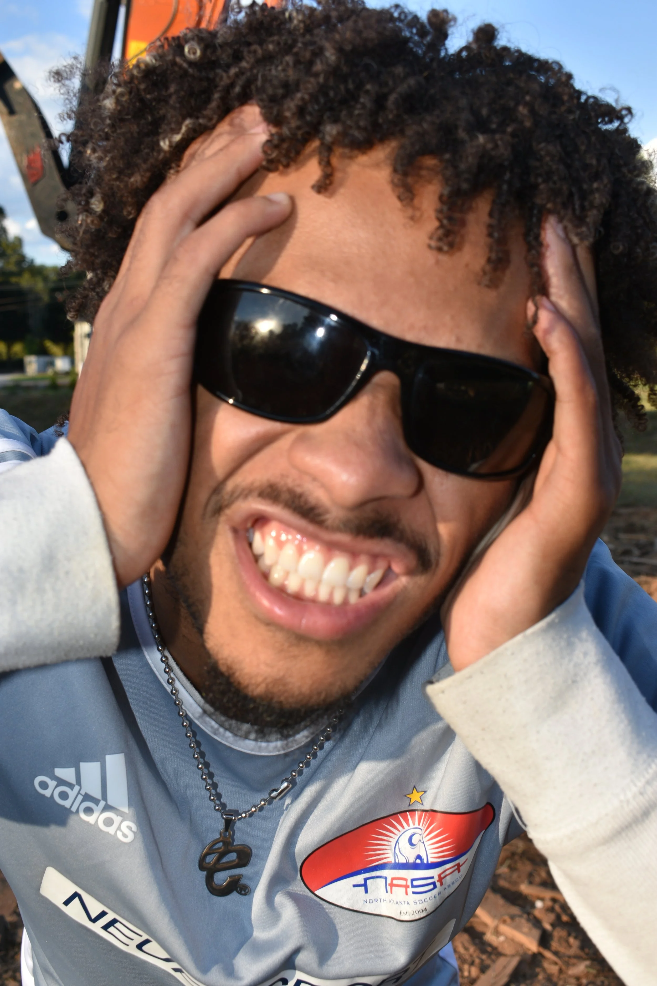 A young man wearing sunglasses, smiling widely, holding his head with both hands, outdoors during daytime, wearing a North Atlanta Soccer Alliance jersey.